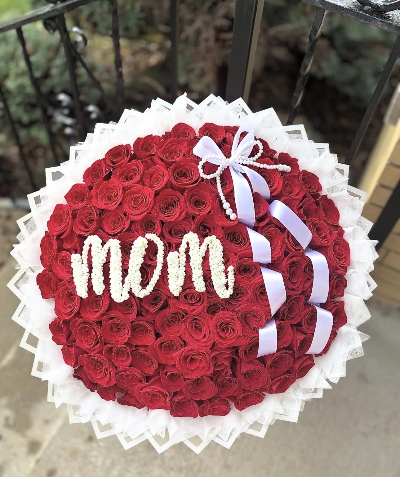 Mom’s RAMO BUCHON - A lush, tightly packed round bouquet of red roses crafted to celebrate Mom with bold, romantic flair-the perfect Mother's Day bouquet or special-occasion arrangement to show appreciation. The large, classic dome of deep red roses is framed by crisp white decorative wrap and finished with a white ribbon and beaded bow, creating a polished, gift-ready presentation that immediately reads as heartfelt and celebratory.  This arrangement features mostly red roses arranged in a compact, buchon-style round shape, accented by white floral lettering that spells "MOM" across the front for a personalized touch. Pink and white tones subtly play against the dominant red to soften the palette and add depth. The white decorative wrap and beaded ribbon lend an elegant contrast and make the bouquet easy to carry and display.  Ideal for Mother's Day, a birthday, or to say "thank you" in a way that will make her feel truly seen, this bouquet feels both romantic and warmly maternal. Because of its tight, structured design, the roses hold their shape beautifully-order in advance to ensure availability and consider pairing with a vase if you'd like longer display life; simple care is to trim stems and refresh water daily to keep the roses looking their best.