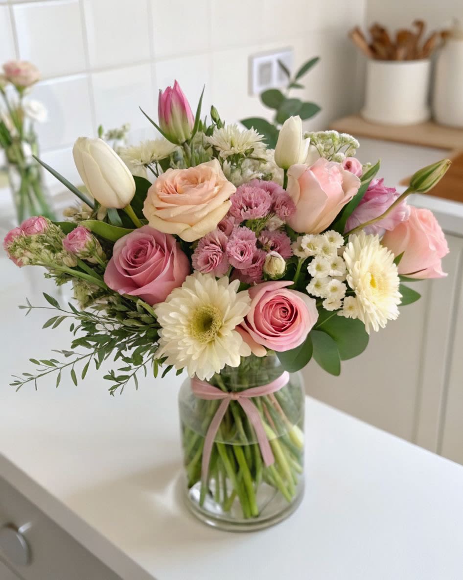 Soft spring vibe  - A gentle palette of blush roses, peach tulips, and soft white daisies creates the Soft spring vibe flower arrangement - a cheerful everyday bouquet perfect for birthdays, anniversaries, or Mother's Day. The mix of roses, tulips, gerbera-like daisies and clustered dianthus gives a romantic, garden-fresh impression that reads as warm and thoughtful to the person you're celebrating.  Set in a clear glass vase tied with a pale pink ribbon, this rounded composition pairs airy greenery with full, textured blooms for a balanced, full look. Pink, white, green, and peach tones blend so the recipient will notice both the lush color and delicate scent; the included chrysanthemums and smaller filler flowers add lasting volume and visual interest.  Send this to make someone feel genuinely seen and cherished - ideal for moms, friends, or a loved one marking a special day or just because. The arrangement arrives ready to display in its vase; refresh water every few days and trim stems slightly to extend bloom life.