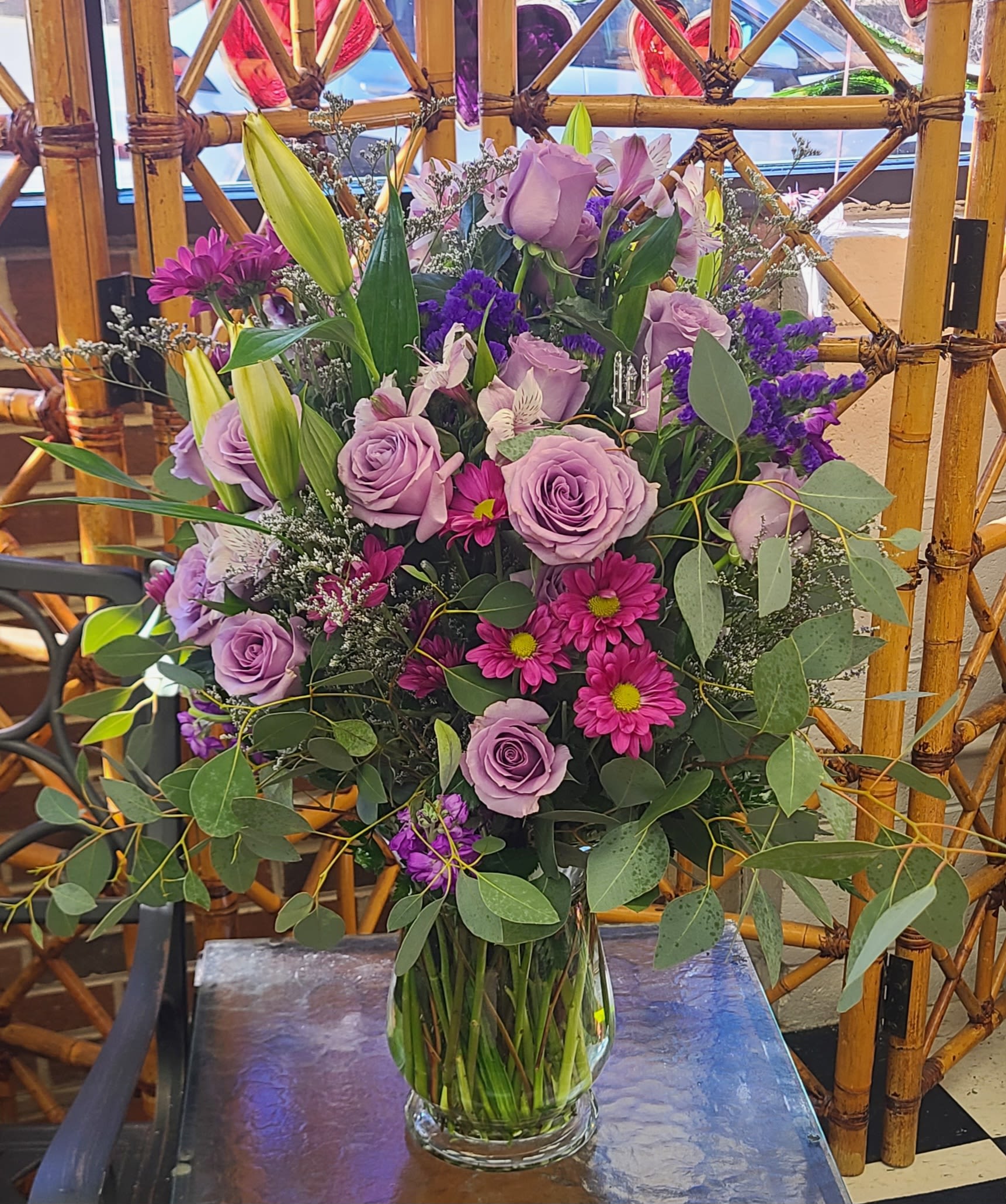 Purple Rose Arangement - Lavender roses blended with pale lily buds and bright magenta daisy mums give this flower arrangement a soft-yet-vivid look that feels both romantic and joyful - perfect as a birthday bouquet, anniversary arrangement, or a just-because surprise for someone you want to celebrate. The first impression is the layered lilac and pink palette that reads like a spring garden: calming lavender tones warmed by pops of magenta and purple accents so the recipient will feel genuinely seen and cherished.  This full, upright design is hand-arranged in a clear glass vase so the long stems are visible and the piece keeps a fresh, natural vibe. It includes roses, lilies, daisy-like mums, alstroemeria and airy white filler, finished with broad, eucalyptus-like foliage that extends outward for a loose, garden-style silhouette. The size and abundant layering make it a substantial gift that looks great on a dining table or bedside - simple care is keeping the vase topped with fresh water and trimming stems if needed to keep the bouquet looking lush.