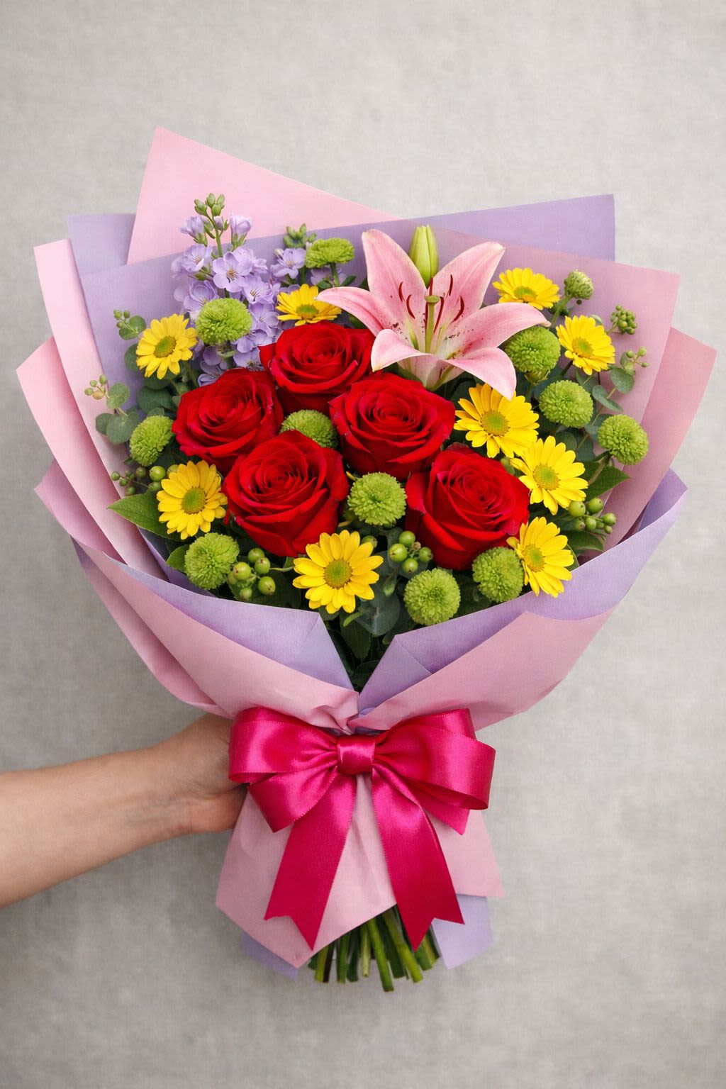 Blush of Love - Five vivid red roses take center stage in this everyday hand-tied bouquet, paired with a single pink lily and cheerful yellow daisy-like blooms to create a lively "Blush of Love" arrangement perfect for anniversaries, birthdays, or simply saying you care. The mix of red, pink, yellow and touches of purple and green delivers a romantic yet joyful message-the kind of bouquet that makes the person you're thinking of feel seen and celebrated.  The bouquet includes roses, a lone pink lily, daisy-style flowers and small green pom-pom accents, all wrapped in layered pastel pink and lavender decorative paper and finished with a bright fuchsia satin ribbon for a polished gift presentation. Textures range from velvety rose petals to the soft, open lily and playful pom-poms, offering visual contrast and a pleasing hand-held size suitable for delivery or pickup.  Ideal as a cheerful personal gesture or celebratory gift, this arrangement arrives ready to present and brightens a bedside table, desk or kitchen counter. For best enjoyment, trim stems and place in fresh water with floral food upon arrival; sizing and specific stem counts reflect the composition pictured, ensuring the bold red roses remain the focal point.