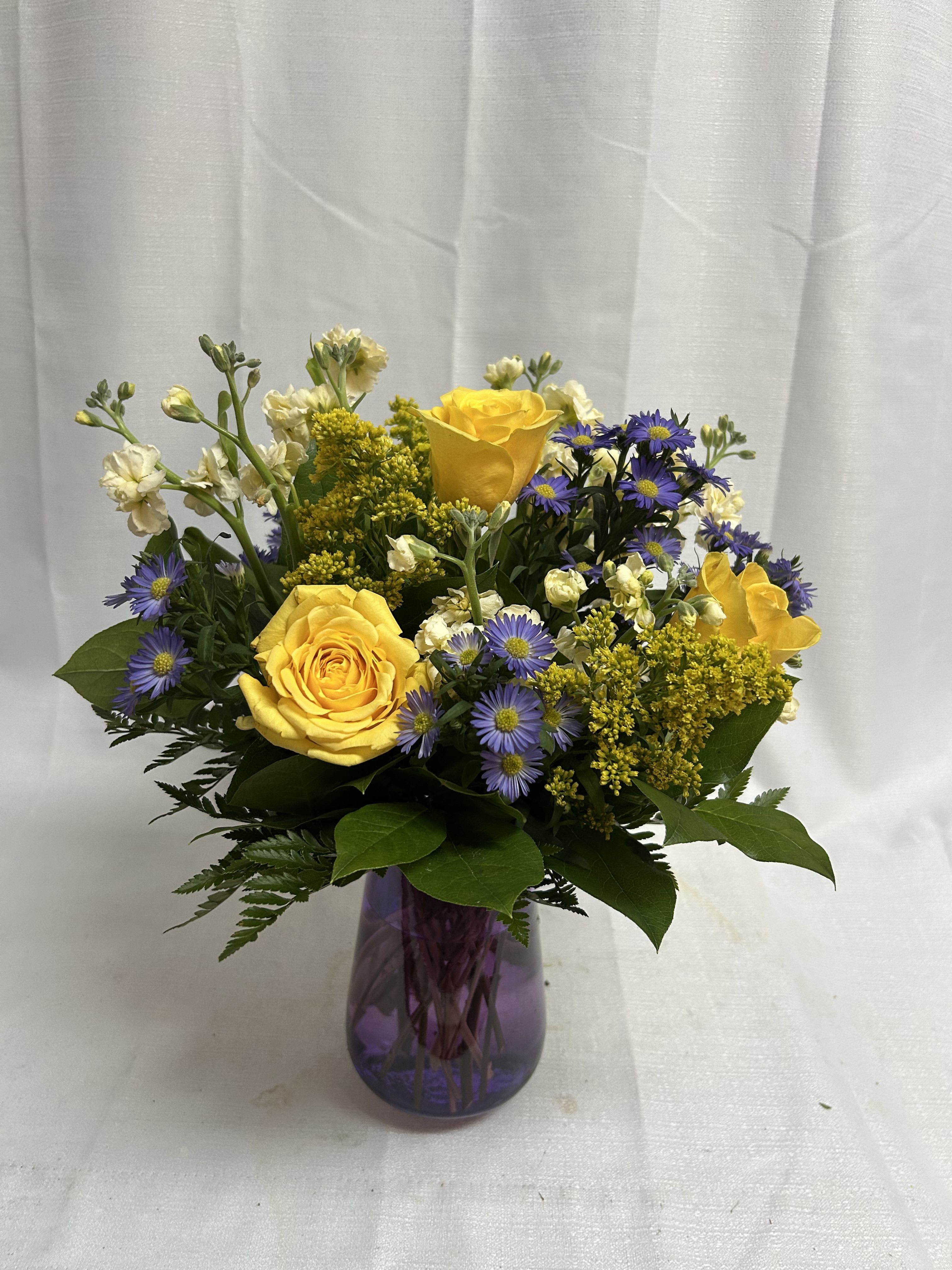 Yellow roses and purple daisies in a purple glass vase