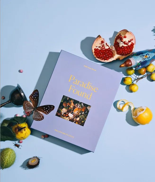 Paradise Found jigsaw puzzle box styled with colorful fruits, insects, and a metal goblet on a blue surface