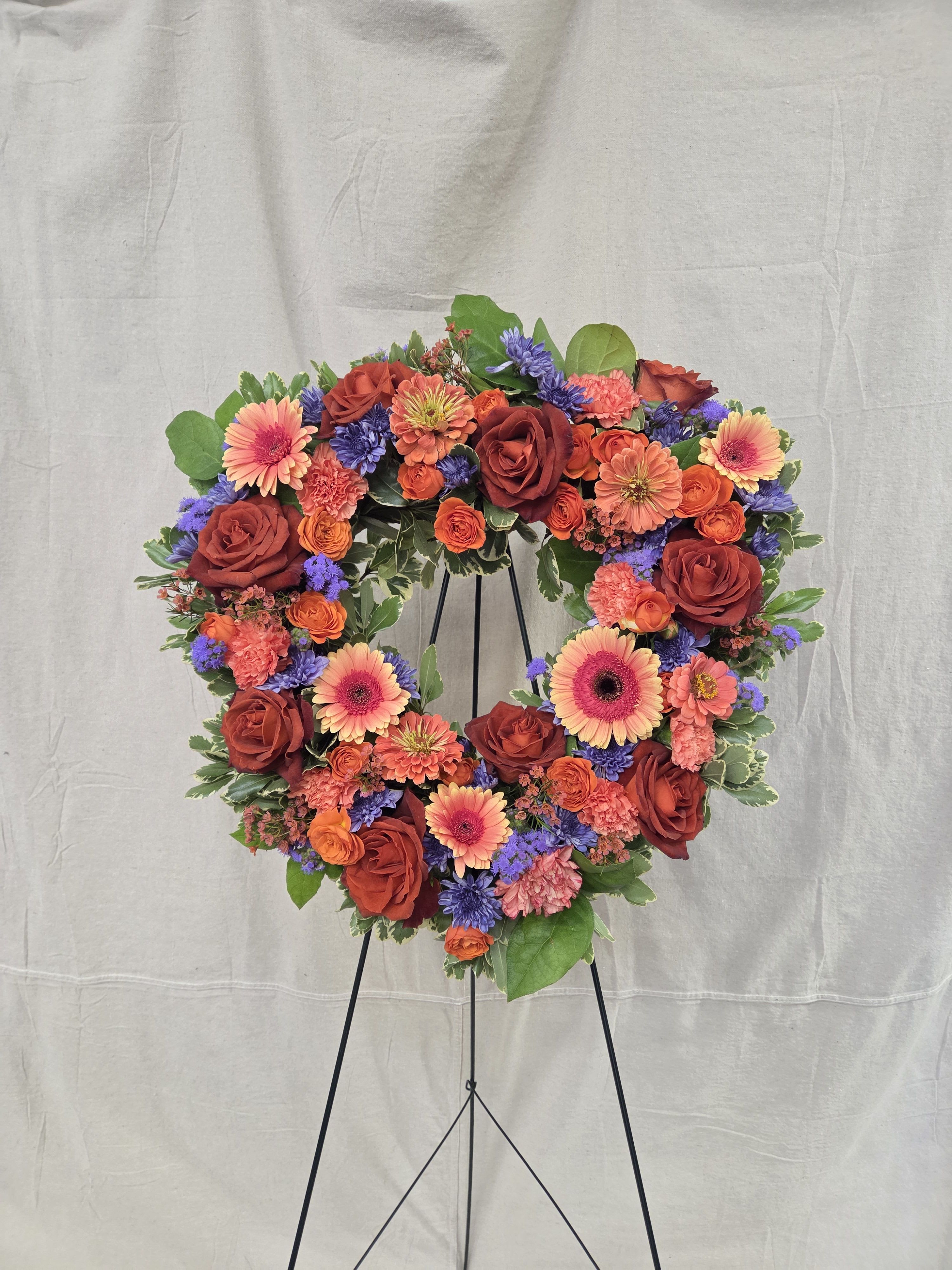 Heart-shaped standing wreath with red roses, orange gerbera daisies, and purple flowers on a black easel