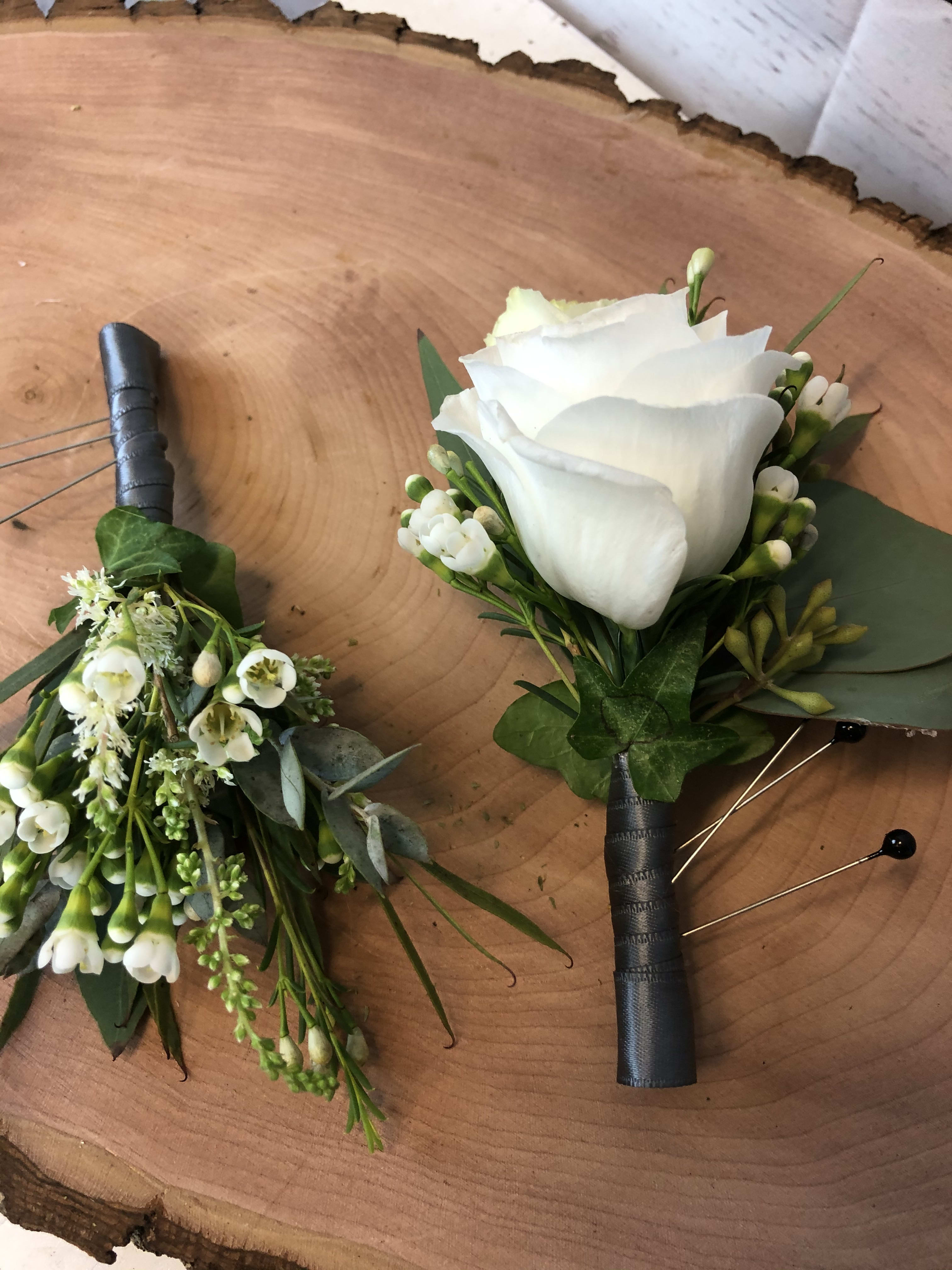 Two white floral boutonnieres with black wrapped stems on a wood surface.