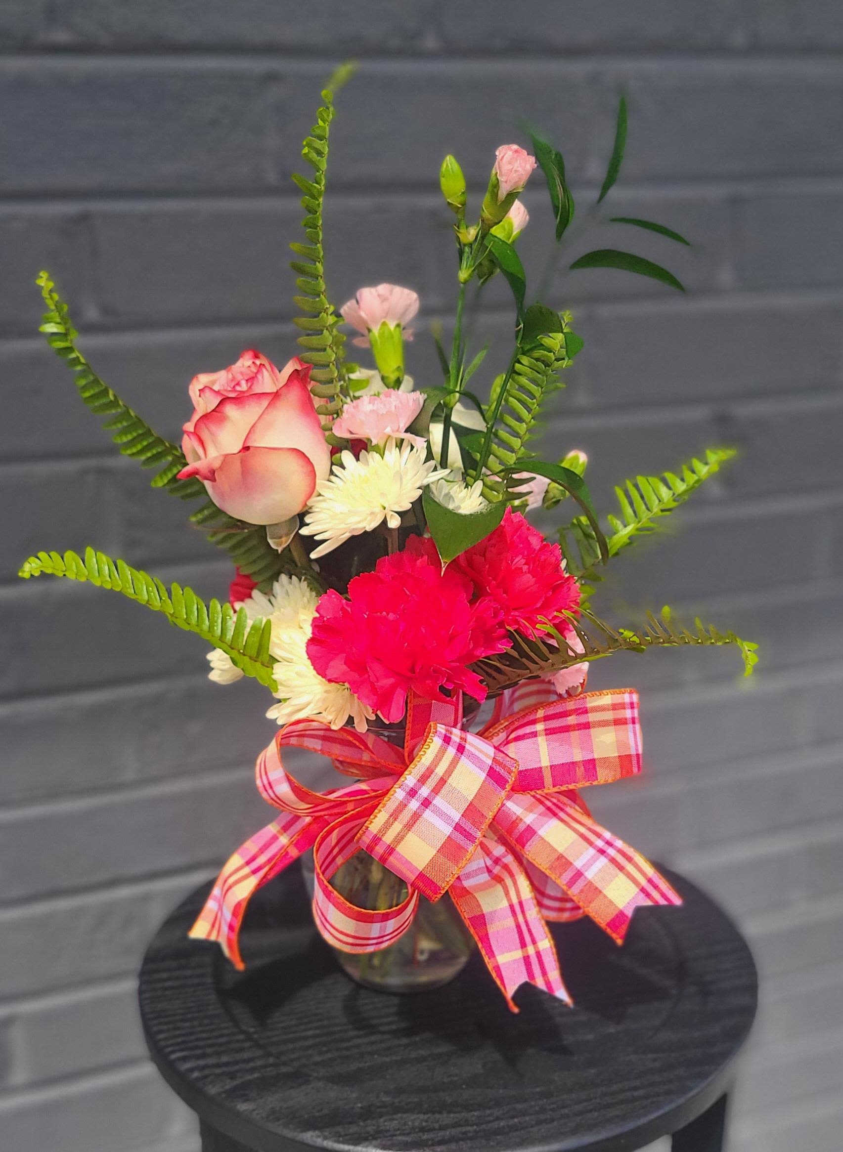 Just Because - Pink-edged roses paired with bright pink carnations and cheerful white daisies make this just-because bouquet feel like a warm surprise from the garden-perfect as a birthday bouquet, a thank-you gift, or an everyday just-because arrangement. Tall fern fronds and slender greenery lift the design into a loose, airy silhouette so the person you're sending it to will feel noticed and genuinely celebrated the moment it arrives.  Arranged in a clear glass vase with a black round base and finished with a large pink-and-yellow plaid ribbon, this mixed pink, white, and green bouquet balances soft romance with playful color. You'll find pale pink spray blooms, small white chrysanthemum accents, and a variety of foliage that add texture and movement; the piece reads as lively and handcrafted rather than overly formal. The vase size and ribbon make it easy to display on a kitchen counter or office desk, and simple care-fresh water and a cool spot away from direct sun-keeps the stems looking fresh for days.