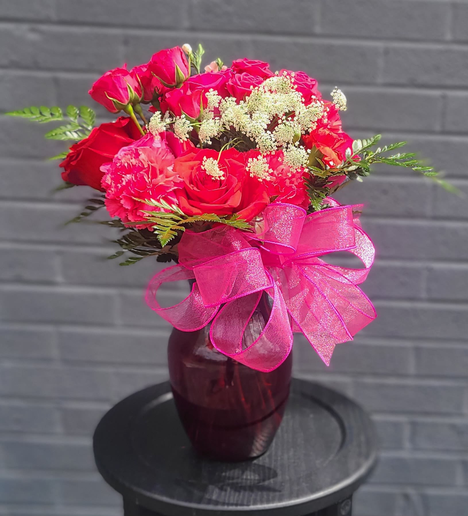 For the Love of It - Vivid pink and red roses clustered with soft pink carnations make an immediate, joyful statement in this birthday bouquet and anniversary arrangement meant for "the one you adore." The compact, rounded shape and saturated hues read as both romantic and celebratory, so it's perfect for anniversaries, Valentine's gestures, birthdays, or a just-because surprise that says you're thinking of them.  Set in a deep burgundy glass vase and finished with a large sheer pink organza ribbon, the design pairs glossy rose petals with airy white filler blooms for contrast and texture. The piece is compact and hand-gifted in feel, with a neat, rounded profile that sits beautifully on a table or desk and presents well straight from the vase.  For the person you want to make feel seen and cherished, this arrangement sends a cheerful, affectionate message without being over the top. Designed as an everyday floral gift, it arrives ready to display-no extra arranging required-so your recipient can enjoy the color, scent, and thoughtful presentation right away.