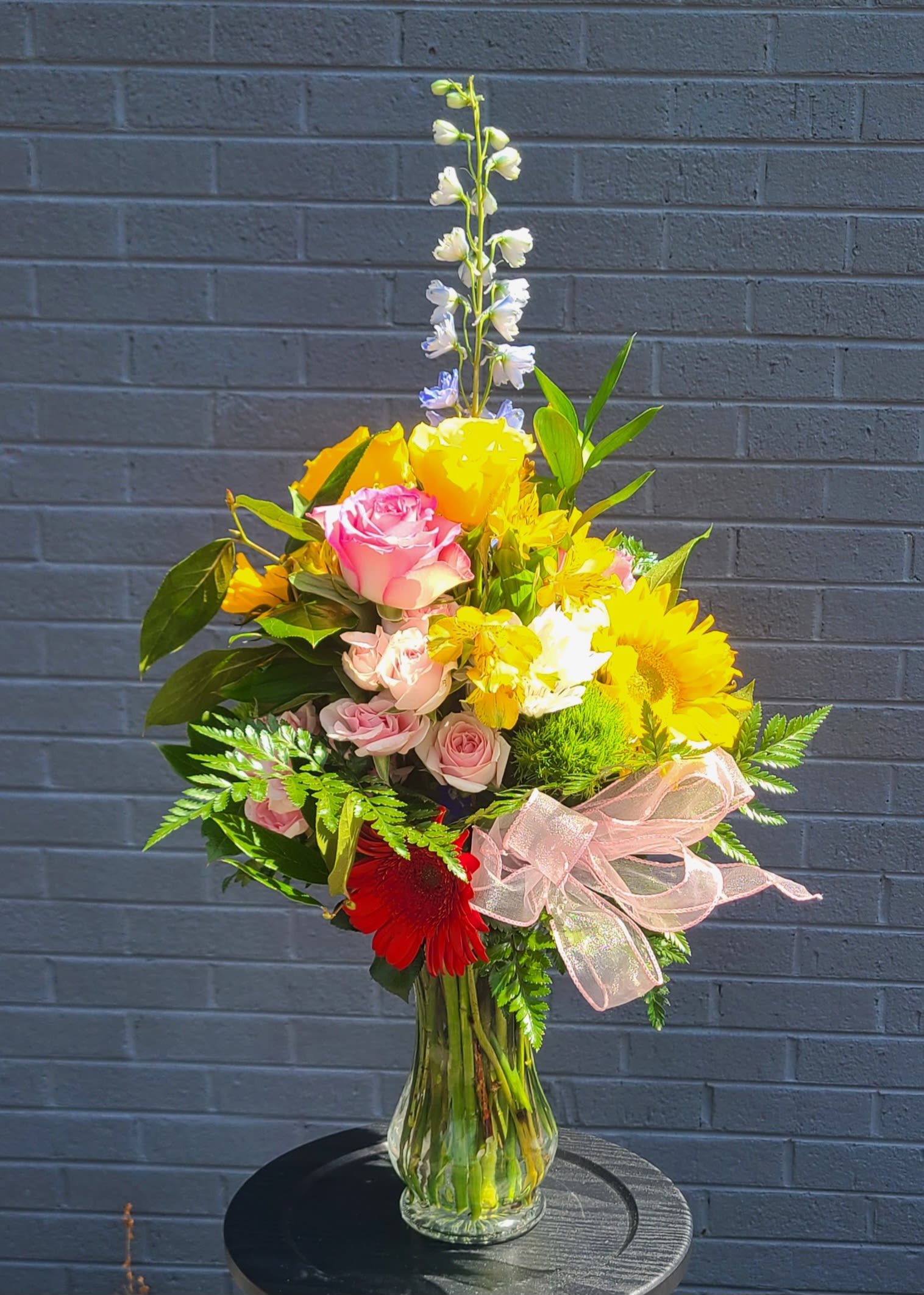 Sunny Days of Joy - Sunshine and soft pastels meet in this tall flower arrangement-perfect as a birthday bouquet, congratulations gift, or a bright "just because" surprise. The design's upward-reaching shape and mix of sunflowers, yellow blooms, pink roses, and a splash of red gerbera create an immediate, joyful impression that will make the person you're thinking of feel celebrated and seen.  Arranged in a clear glass vase and tied with a pink sheer ribbon, the bouquet layers rounded rose heads and large sunflower faces against white accents, pale blue delphinium spikes, alstroemeria, and mixed daisies for texture and movement. A single slender stem rises above the cluster to add elegant height; the color palette of yellow, pink, white, red, and blue keeps the look cheerful and fresh without feeling fussy.  Designed to stand tall on a side table or brighten a mantel, this arrangement arrives polished and ready to gift. The vase presentation and ribbon finish make it an easy choice when you want to send warm, sunny wishes that truly lift someone's day.