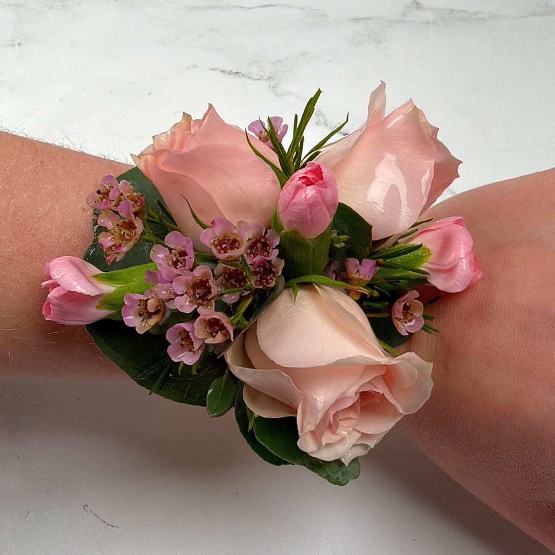 Blushing Rose Corsage - Pale blush roses clustered with pink carnations and tiny pink waxflower fillers create a soft, romantic wrist corsage perfect for prom, weddings, or any special celebration. The Blushing Rose Corsage is a compact floral corsage designed to sit gracefully on the wrist, the color palette of blushes, pinks, white accents and rich green foliage reads delicate and feminine, making the person wearing it feel cherished and elegantly finished.  This wrist corsage features tightly arranged pale pink roses, a bright pink carnation bud for contrast, and small clustered pink filler flowers for texture, all framed by glossy dark green leaves. It's secured on a metallic gold cuff for a dressy, polished look. The compact, rounded design sits neatly on the wrist without overwhelming the wearer's outfit-ideal prom dates or a meaningful gift that says "you're special."