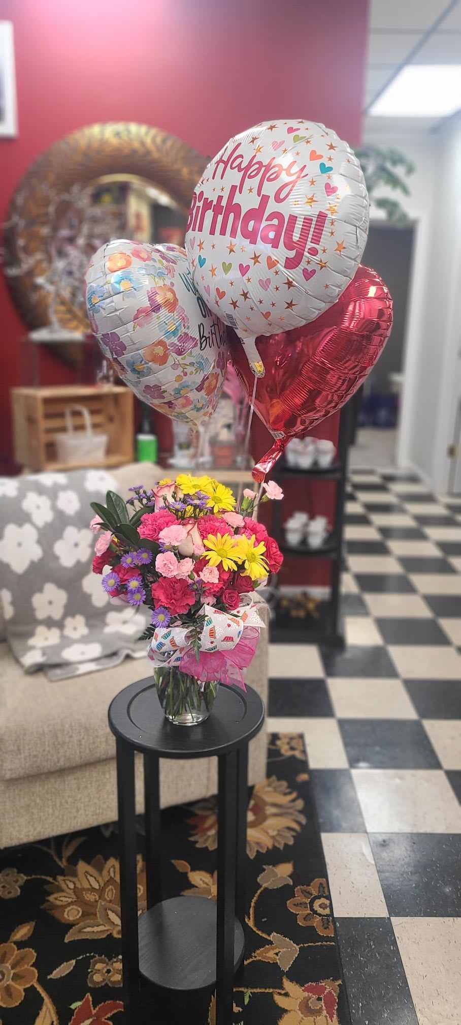 BIG BIRTHDAY!!! - Bright, playful, and unmistakably celebratory - this birthday bouquet brings a burst of pink, yellow, purple, and white blooms topped with three helium birthday foil balloons. Perfect as a birthday bouquet or cheerful surprise for someone turning a year bolder, it's the kind of arrangement that makes the person you're thinking of feel celebrated the moment it arrives.  A generous mixed bouquet of roses, carnations, and daisy-like blooms is arranged in a clear glass vase for a layered, rounded silhouette that reads lively from every angle. A patterned decorative ribbon and ribbon bow finish the vase for a gift-ready presentation; the dense clusters of small rose heads and carnations give the arrangement a soft, textured look while the bright yellow and purple accents keep the palette energetic.  Ideal for birthdays, congratulations, or any cheerful occasion where you want to brighten someone's day, this arrangement arrives ready to display - balloons already attached and the vase filled so the recipient can enjoy it immediately. For best long-lasting color and freshness, place in a cool spot away from direct sun and refresh the water every couple of days.