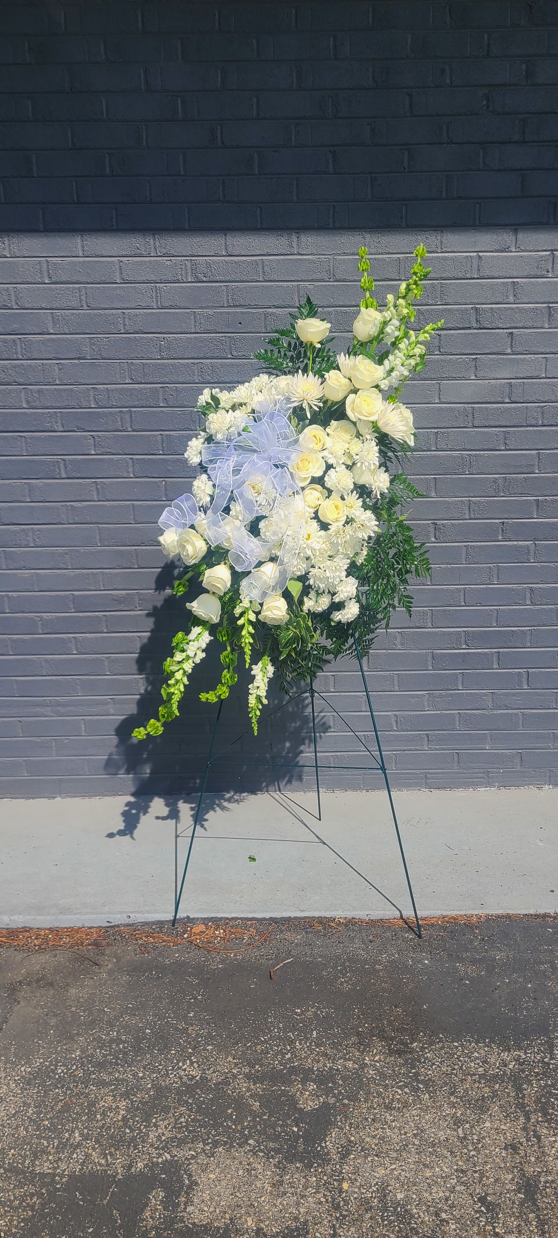 Standing Spray Pure White - Pure white roses and clusters of daisy-like chrysanthemums form a serene, vertical tribute in this standing spray-perfect for a sympathy arrangement or memorial service. Tall snapdragon spikes and soft green foliage rise above the main body to create a formal, upright silhouette that reads as both respectful and refined, while the pale lavender ribbon offers a gentle, consoling touch.  The spray is arranged on a metal floral easel and built for display at services or remembrance settings; its layered shape and compact clusters of white blooms give a full, dignified presence without overpowering the space. White roses, white chrysanthemums, upright snapdragons, and airy greenery combine for a calm white-and-green palette with a hint of lavender from the sheer ribbon.  Designed to honor and comfort, this arrangement is the kind of floral tribute that helps the person you're thinking of feel remembered and respected. The metal easel provides stable presentation at funerals, memorials, or other solemn events-order with confidence for delivery to a service or venue.