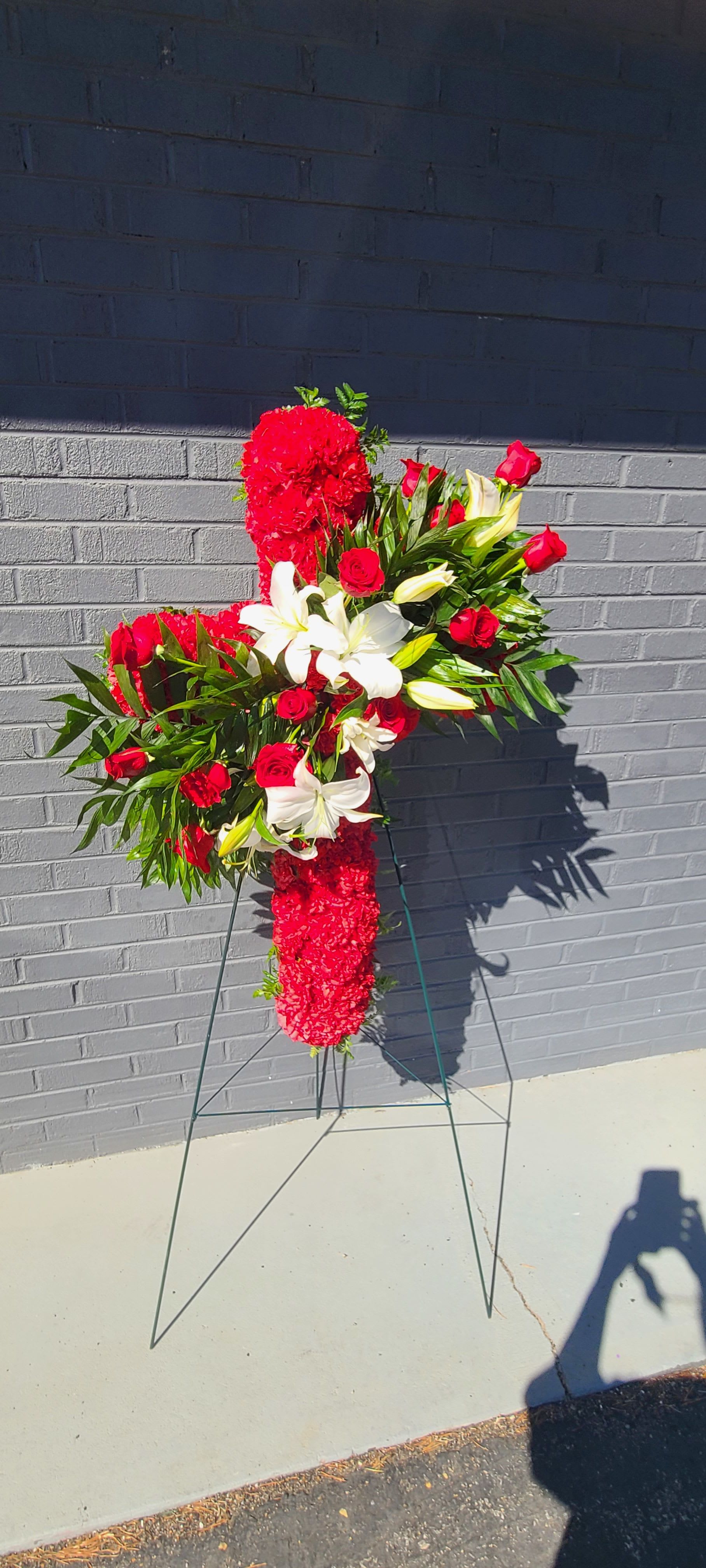 Standing Cross  - A tall, solemn standing cross that speaks with clarity and comfort-this sympathy flower arrangement is crafted for memorial and funeral services to honor a life with dignity. The vertical cross silhouette, mounted on a wire/metal easel, creates an immediate devotional presence so the person you're remembering feels reverence and warmth through color and form.  Dense clusters of red roses and red carnations provide a vivid, heart-centered red, while white lilies and a few unopened lily buds introduce pure, uplifting contrast and gentle fragrance. The design is generously covered with flowers for a formal, high-impact look; the cross shape is substantial and visible from a distance, making it suitable for church services, visitation rooms, or graveside memorials.  Choose this arrangement when you want to express deep sympathy, respect, and heartfelt remembrance-the colors and structure send a clear message of honor. It arrives ready on a sturdy metal/wire easel for immediate display; light care involves keeping the oasis hydrated until the service.
