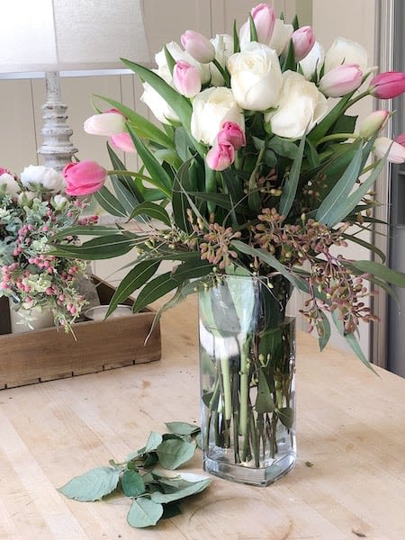 Vase  - A dense cluster of creamy white roses surrounded by soft pink tulips creates an airy, feminine centerpiece that's perfect as a birthday bouquet, anniversary surprise, or get-well gift. This flower arrangement places the recipient at the center - she'll feel gently celebrated and comforted by the cloud of blooms and the tender palette.  The roses form a plush focal core while pink tulips in various stages of opening fan around them, their long stems visible in a tall clear rectangular glass vase for a fresh, garden-cut look. Long, slender foliage and seeded accents give height and a natural texture, and the arrangement arrives set on a wooden tray with a small ceramic container and table lamp nearby in the styling to suggest how it livens a bedside or coffee table. Colors are soft white and blush pink for an elegant, versatile presentation.  Perfect for brightening a home or bedside, this arrangement fits well on dining tables, mantels, or next to a reading chair and makes a thoughtful everyday gift as well as a tender way to mark anniversaries or recovery. Standard sizing keeps the composition full and easy to display; replace water every 2-3 days and trim stems slightly to extend vase life.