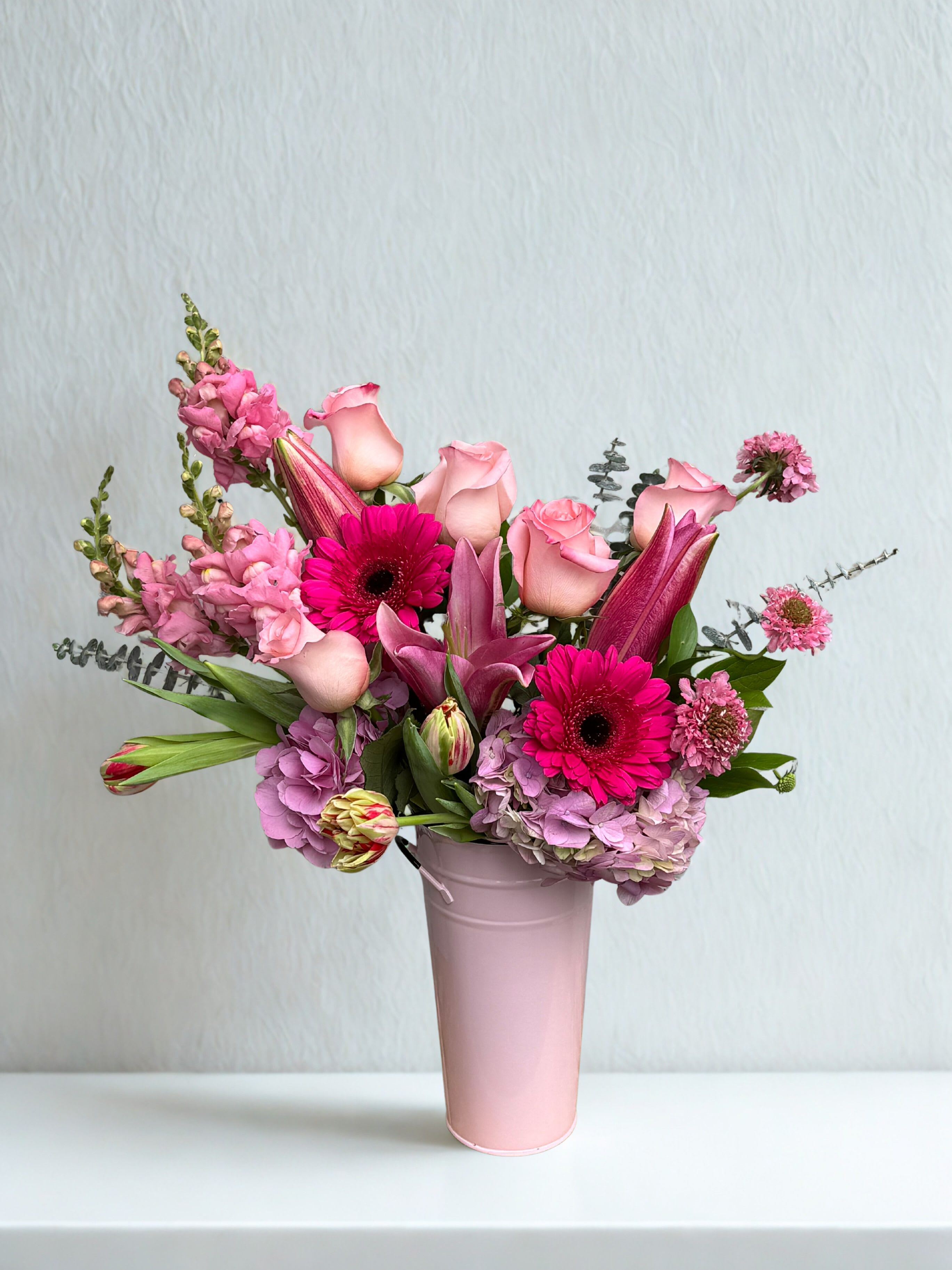 Lovely Lolly - A vibrant mix of pink blooms featuring roses, gerber daisies, lilies and hydrangea accented with fresh greens and arranged in a soft pink metal vase. 