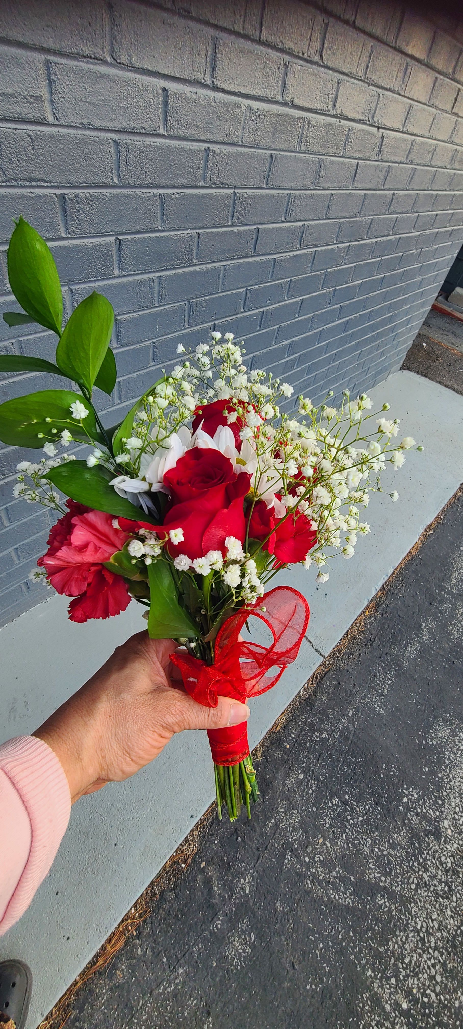 Custom Bouquet - Vivid red roses take center stage in this handheld flower arrangement, making it an instantly romantic choice for an anniversary, Valentine's Day, or a heartfelt "I'm thinking of you" surprise. The classic red-and-white palette-bright roses paired with white daisies or airy baby's breath and touches of soft pink-creates a timeless, celebratory look that reads as both passionate and sincere to the person receiving it.  This compact, slightly loose hand-tied bouquet mixes roses, carnations, daisies, and filler blooms with glossy green foliage for height and contrast. Each bouquet is gathered tightly at the stems and finished with a red ribbon wrap for a gift-ready presentation; textures range from the smooth rose petals to the feathery baby's breath for an inviting, natural feel. The size is portable and perfect for hand-delivering, making it ideal for birthdays, thank-you gestures, or everyday expressions of love.