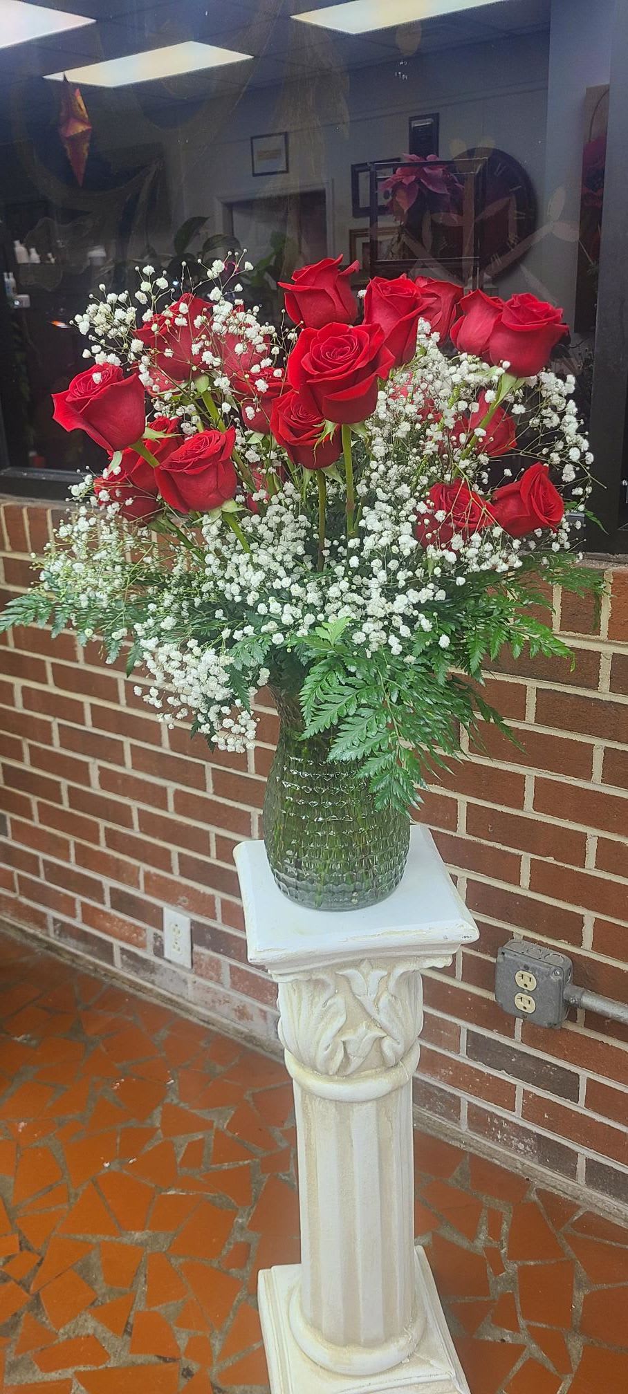 Pure Love - A dense cluster of velvety red roses crowned by airy sprays of white baby's breath gives this flower arrangement a timeless, romantic presence-perfect as a birthday bouquet for a sweetheart, an anniversary roses gift, or a Valentine's Day surprise. The strong red-and-white contrast and the arrangement's upright, formal silhouette also make it an appropriate choice for sympathy or funeral use when you need a respectful, classic expression.  The roses are gathered tightly above a cloud of delicate white baby's breath and set in a clear textured glass vase (some images show a green-tinted textured vase variant) that sits on a decorative white pedestal stand for added height and ceremony. The composition reads full and substantial while remaining refined: rich red blooms, crisp white fillers, and green foliage provide striking color contrast and pleasing texture.  Give this to the person you want to make feel deeply seen and loved-whether marking a milestone, saying "I love you," or offering comfort. Ships ready to display on its pedestal stand; please follow standard floral care of fresh water and a cool spot to extend vase life.