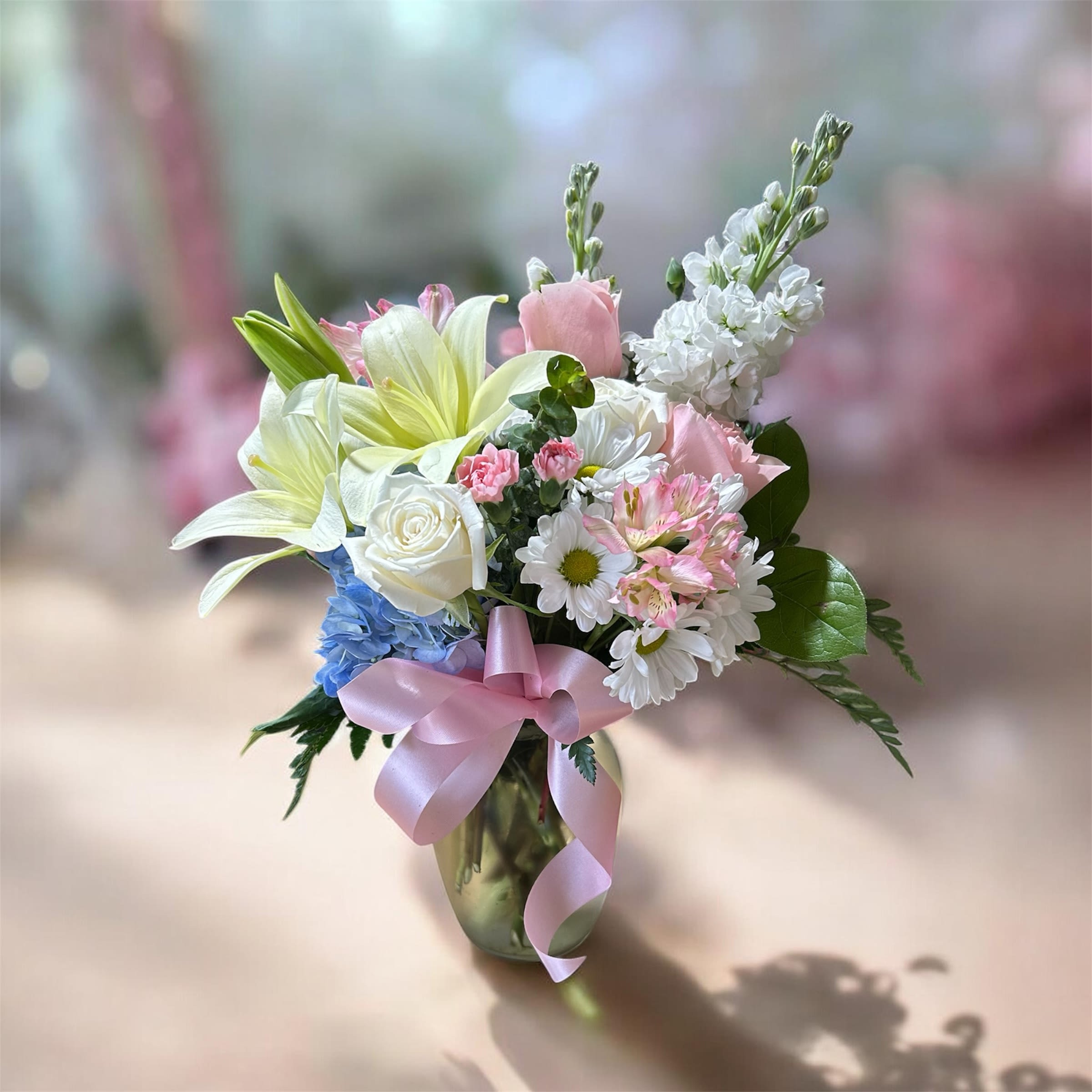     Timeless Bloom - Pale yellow-white lilies and creamy white roses mingle with pink carnations and alstroemeria in a soft, romantic spray-Timeless Bloom is a spring-worthy flower arrangement perfect for a birthday bouquet or a tender celebration. The rounded, full composition reads cheerful and feminine, with a cluster of blue blooms adding a gentle, unexpected pop and tall white stock rising above the main cluster to give graceful height and movement that will make the recipient feel truly seen.  Arranged in a clear glass vase and tied with a pink satin ribbon, the bouquet pairs delicate pastels-white, pink, blue, and fresh green foliage-for an elegant, layered texture that smells subtly sweet and feels luxurious to hold. Flowers include lilies, roses, daisies, carnations, alstroemeria, and stock, presented in a compact, rounded shape ideal for tabletops and bedside tables.  Perfect for celebrating a friend, mother, or someone special on a spring birthday, new baby visit, or to say "I'm thinking of you," Timeless Bloom arrives gift-ready in the vase with bow. For best enjoyment, keep the vase filled with fresh water, trim stems occasionally, and place away from direct heat to extend bloom life.