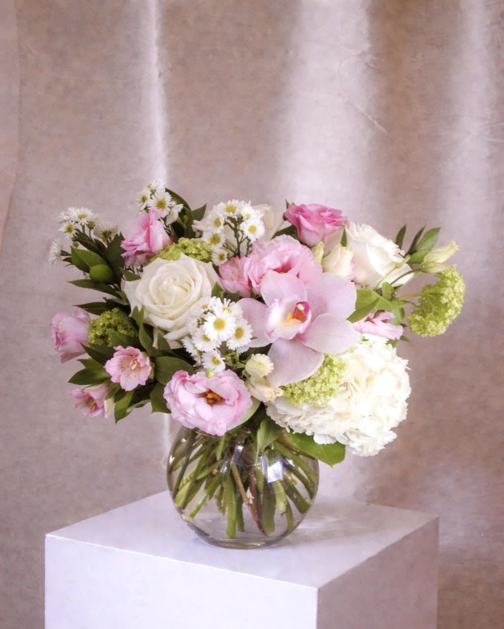 Sweet Mama - A soft, garden-style arrangement in blush and white tones, featuring roses, lisianthus, delicate asters, hydrangea, and a focal pink orchid. Designed in a clear glass vase with visible stems, it has an airy, freshly gathered feel—light &amp; feminine.