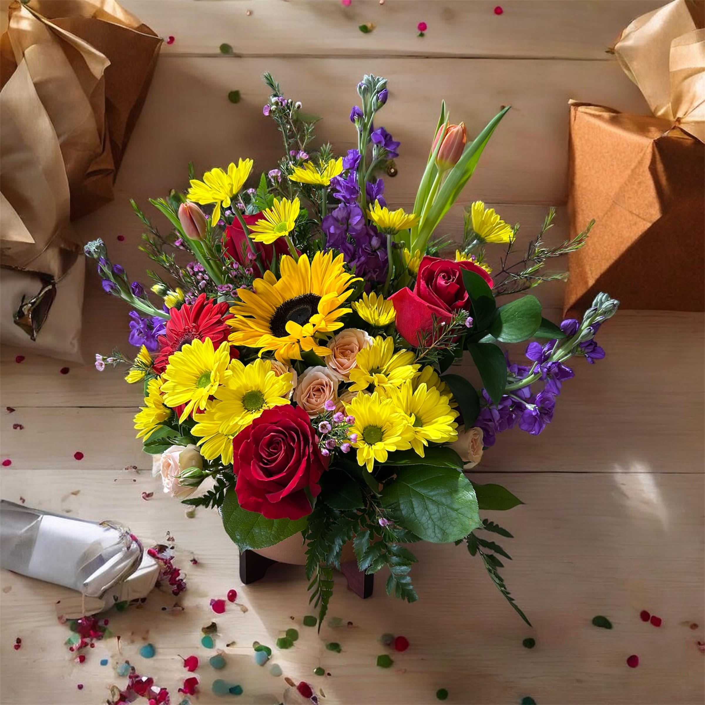     Sunburst Surprise - A joyful burst of color greets the recipient with Sunburst Surprise, a celebratory flower arrangement perfect for birthdays, congratulations, or a spring celebration. This rounded bouquet reads bright and festive-designed to make someone feel truly seen-mixing cheerful yellow daisies and yellow accents with soft white hydrangea clusters and pink-and-white lilies for immediate visual lift. Deep red and peach roses tucked into the mix add a warm, slightly romantic note for a loved one, while purple chrysanthemum-like blooms and snapdragons bring contrast and texture.  The design pairs small blue daisy-like flowers and alstroemeria with lush glossy foliage for depth, arranged in a clear glass vase tied with a wide purple ribbon for a polished gift presentation. The bouquet's low-box moments and denser vase option (as shown) create a full, rounded silhouette about the size of a generous centerpiece-bright, tactile, and layered with varied petal shapes and scents. Packaged with a ribboned gift box, Sunburst Surprise sends a cheerful, confident message: the person you're thinking of will feel celebrated, loved, and uplifted.