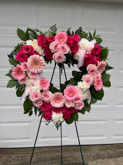 Heartfelt Condolences  - A heart-shaped wreath built from lush clusters of pink and white roses, pink gerbera daisies, and white carnations creates an elegant, ceremonial tribute for sympathy and funeral services. The arrangement's full, symmetrical silhouette on a black metal easel feels both formal and tender-the kind of floral expression that lets the person you're honoring feel genuinely remembered and cherished.  Bright magenta accents rim the outer edge while softer blush tones nestle toward the center, all set against broad, dark green foliage that gives the piece structure and depth. Finished with a delicate lace ribbon, the wreath reads as romantic and reverent at once; the dense placement of blooms ensures a continuous field of color and texture from every viewpoint.  Appropriate for a memorial service or to bring comfort to grieving loved ones, this heart-shaped design sends a clear message of care and remembrance. The easel stand and lace ribbon arrive ready for display; for delivery or placement timing, please select your service options at checkout so the tribute arrives fresh and on schedule.