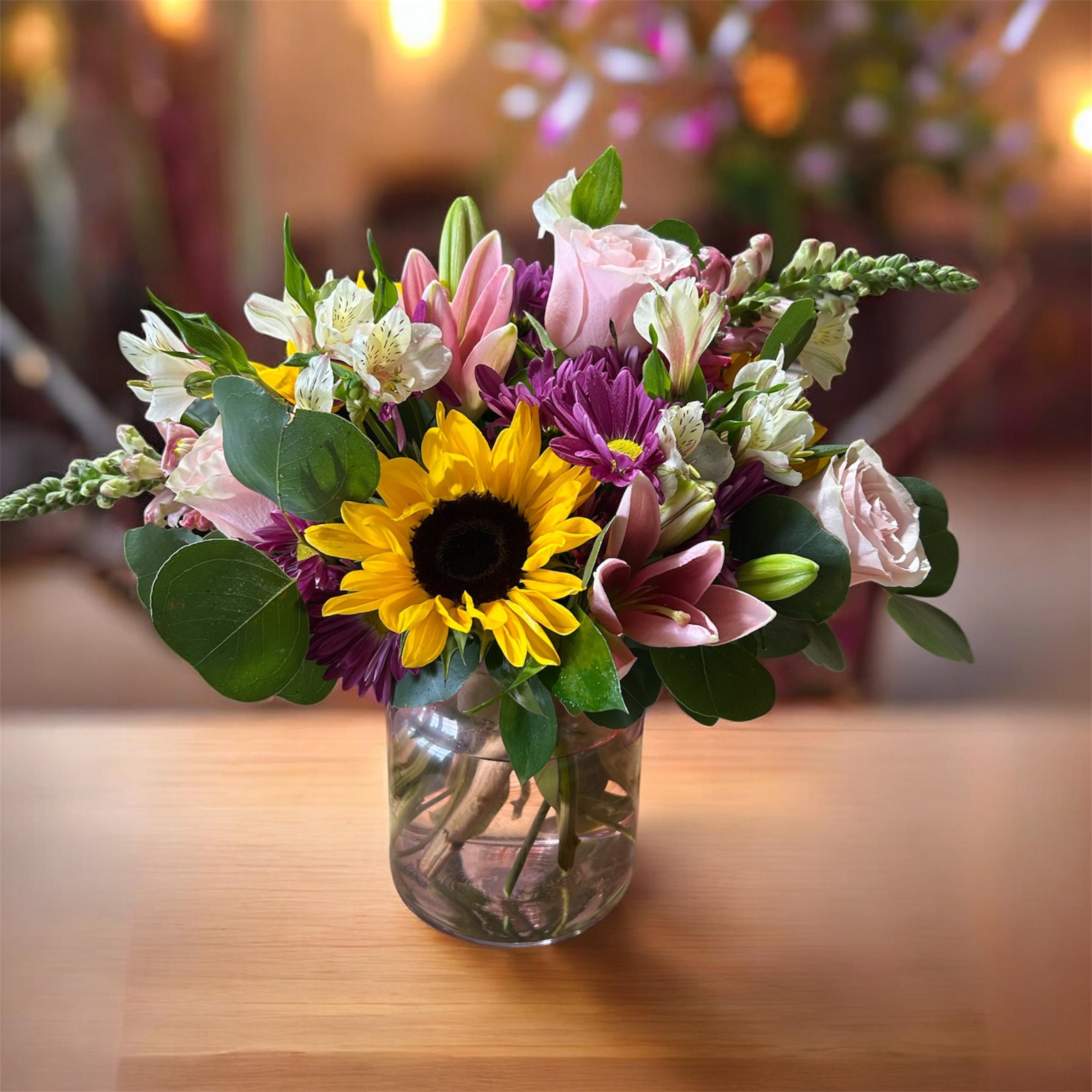 Sunset Garden - A bright sunflower anchors this mixed-color flower arrangement, a cheerful garden-style bouquet perfect as a birthday bouquet, thank-you gift, or spring celebration centerpiece. The warm yellow sunflower paired with soft pink roses, fragrant lilies, white alstroemeria, and purple daisy-like chrysanthemums creates an upbeat, welcoming impression that will make the person you're thinking of feel genuinely celebrated.  Arranged in a clear glass vase, the composition has a full, rounded, slightly informal hand-tied look that reads fresh and casual on a table or entry console. Expect a lively blend of textures-velvety rose petals, bold sunflower faces, starry alstroemeria, trumpet-shaped lilies, and textured chrysanthemum blooms-in a compact, gift-ready size that suits everyday gifting. Care: change the water every other day, trim stems at an angle, and keep away from direct heat to extend vase life.