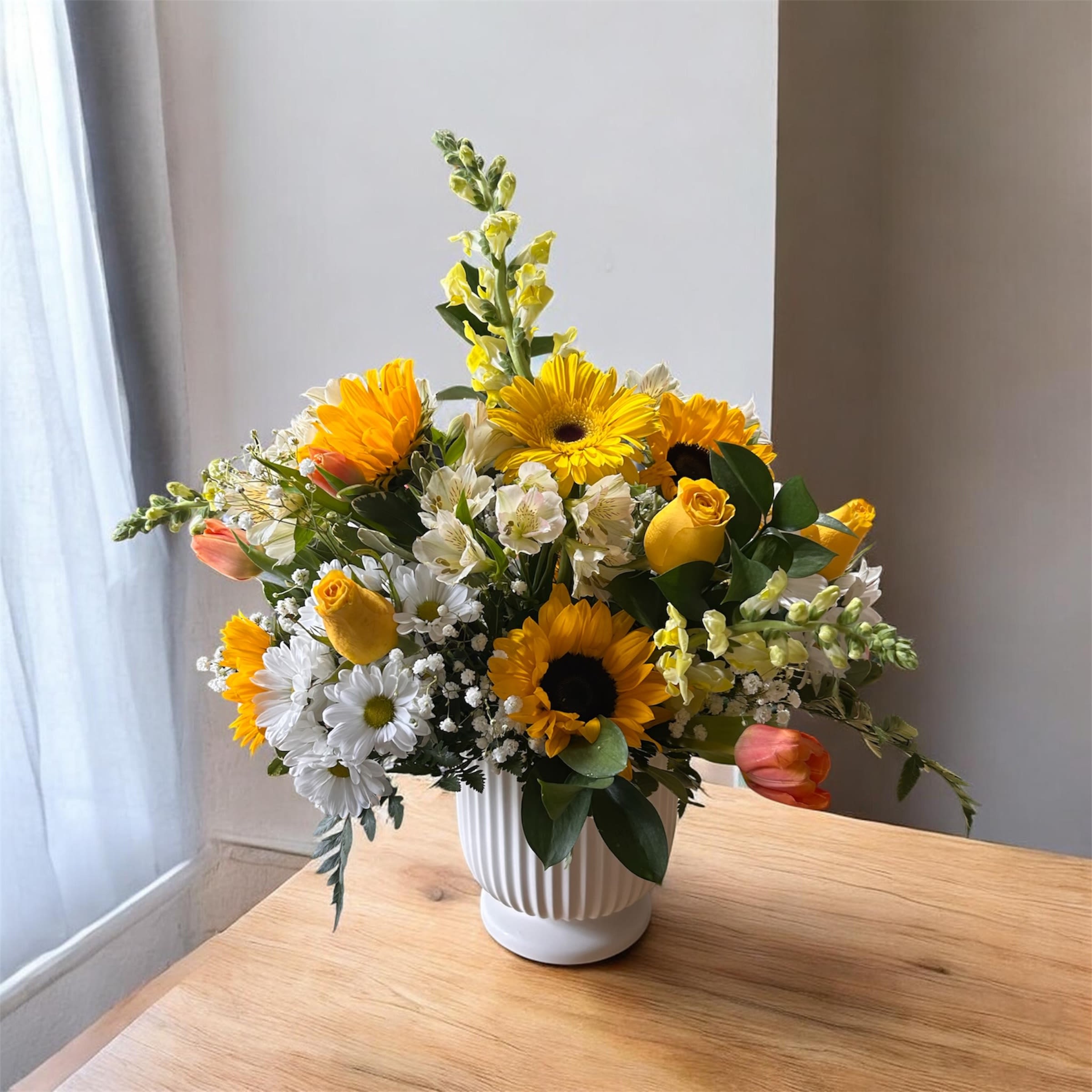 Fresh Start Bouquet - Sunflowers and cheerful daisies take center stage in this Fresh Start bouquet, a bright flower arrangement perfect for a birthday bouquet, spring celebration, or a thoughtful "thinking of you" gift. The sunny yellow and crisp white palette-softened by blush pink tulips and pale yellow rose buds-creates an uplifting, garden-fresh impression that will make the person you're sending it to feel genuinely celebrated and cared for.  Arranged in a ribbed white ceramic vase, the mix includes sunflowers, daisy-like blooms, alstroemeria, tulips, snapdragons, and roses, with glossy green foliage and small white filler flowers adding texture and depth. The rounded, full composition reads lively without feeling fussy, offering a mix of heights and shapes that bring movement and a natural, hand-picked look.  Ideal for everyday gifting, get-well wishes, or welcoming spring, this bouquet arrives ready to display in its sturdy white vase. For best enjoyment, keep in fresh water, trim stems every few days, and place away from direct heat or drafts so the blooms stay vibrant as long as possible.