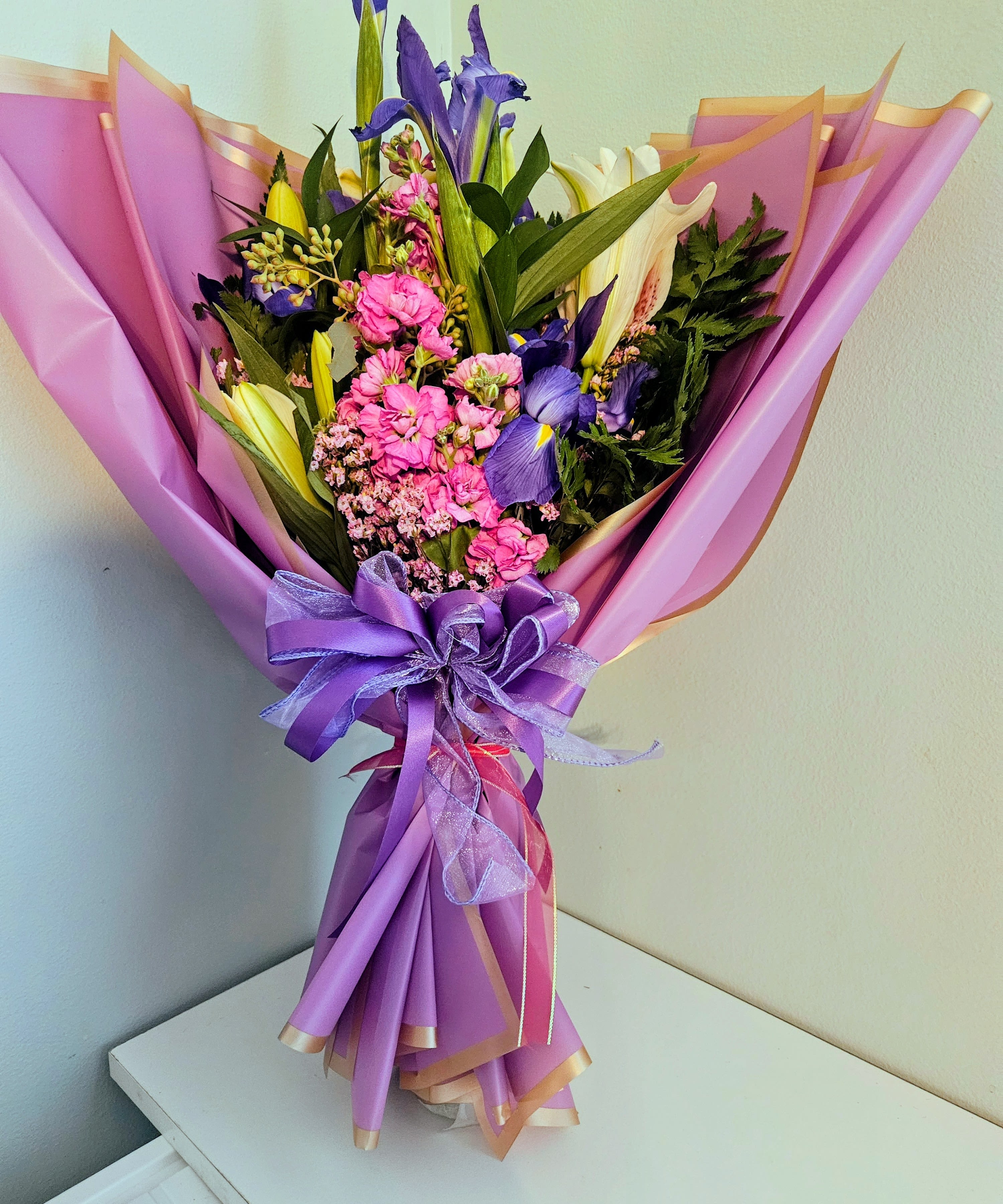 "Amethyst Garden" - Tall purple iris soar above a lush cluster of white lilies and pink carnations in this Amethyst Garden flower arrangement-an everyday bouquet perfect for birthdays, thank-you gifts, or simply to brighten someone's day. The mix of cool purples and bright pinks reads both cheerful and gently romantic, making the person you're thinking of feel celebrated and truly seen.  This hand-tied arrangement layers stately iris, fragrant white lilies, ruffled pink carnations, and delicate filler blooms for texture and movement. Wrapped in lavender decorative paper with a soft peach edge and finished with a large purple satin and sheer ribbon bow, it arrives gift-ready with polished presentation and a lively color story of pink, purple, white, and a touch of sunny yellow.
