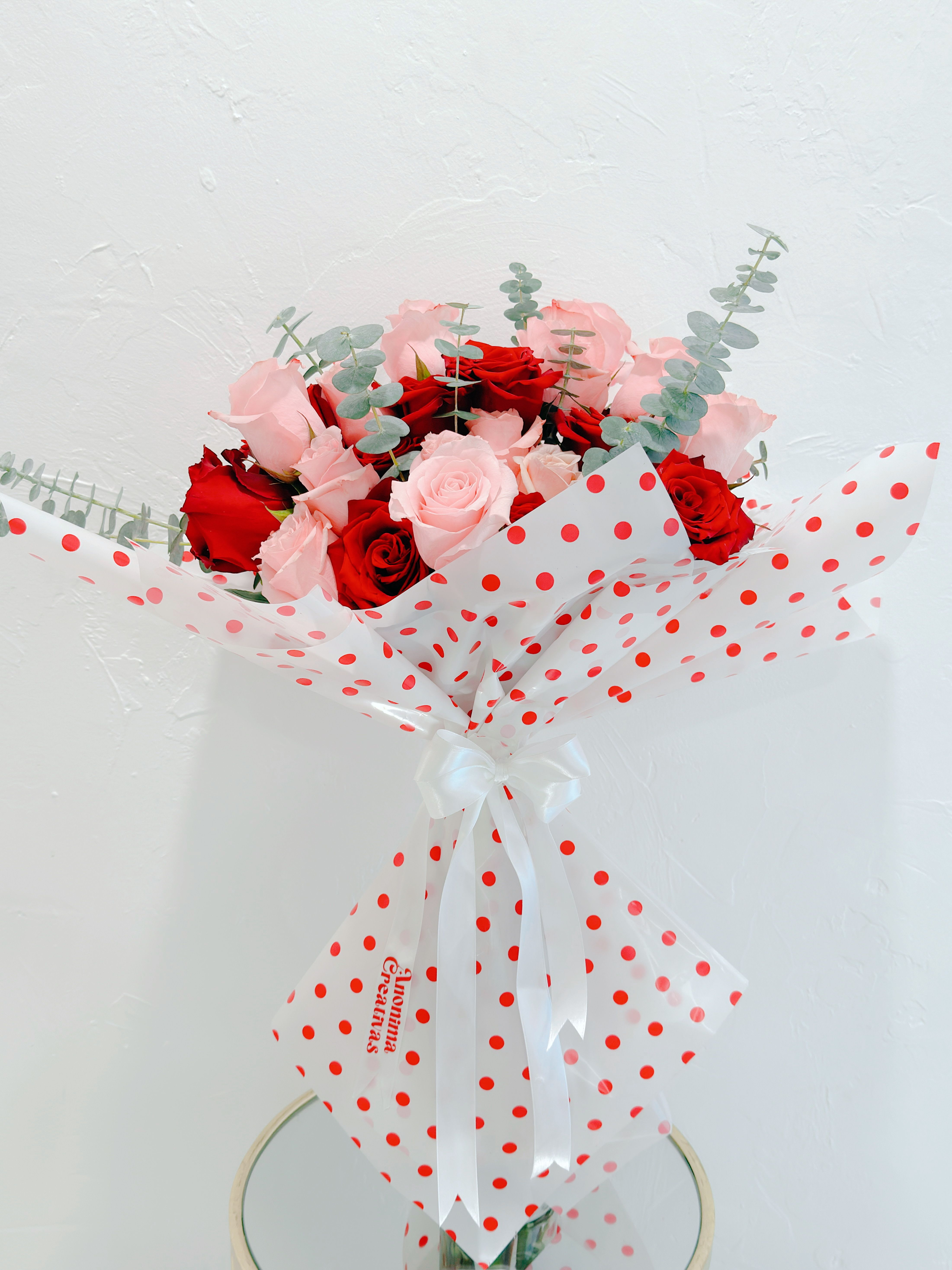 Bouquet Pink  - Elegant and delicate bouquet featuring a beautiful mix of red and pink roses, creating the perfect balance between passion and tenderness. Wrapped in translucent paper with red polka dot details and finished with a satin white bow for a modern, sophisticated touch. Accented with fresh eucalyptus foliage, this arrangement is ideal for special occasions such as birthdays, anniversaries, or Mother’s Day.  A heartfelt gift designed to express love with style and freshness. 