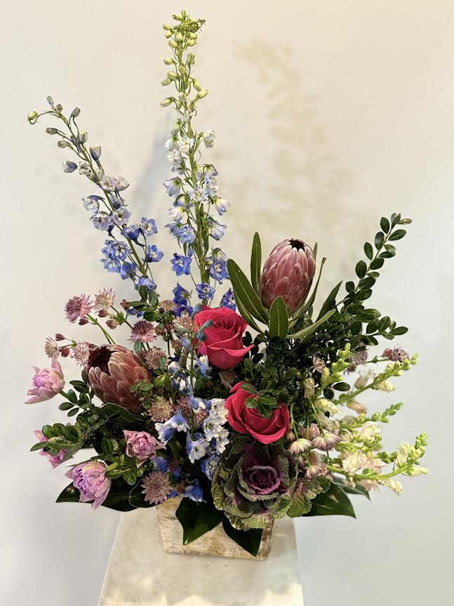 Tall mixed bouquet with pink roses, blue delphinium, and protea in a square vase