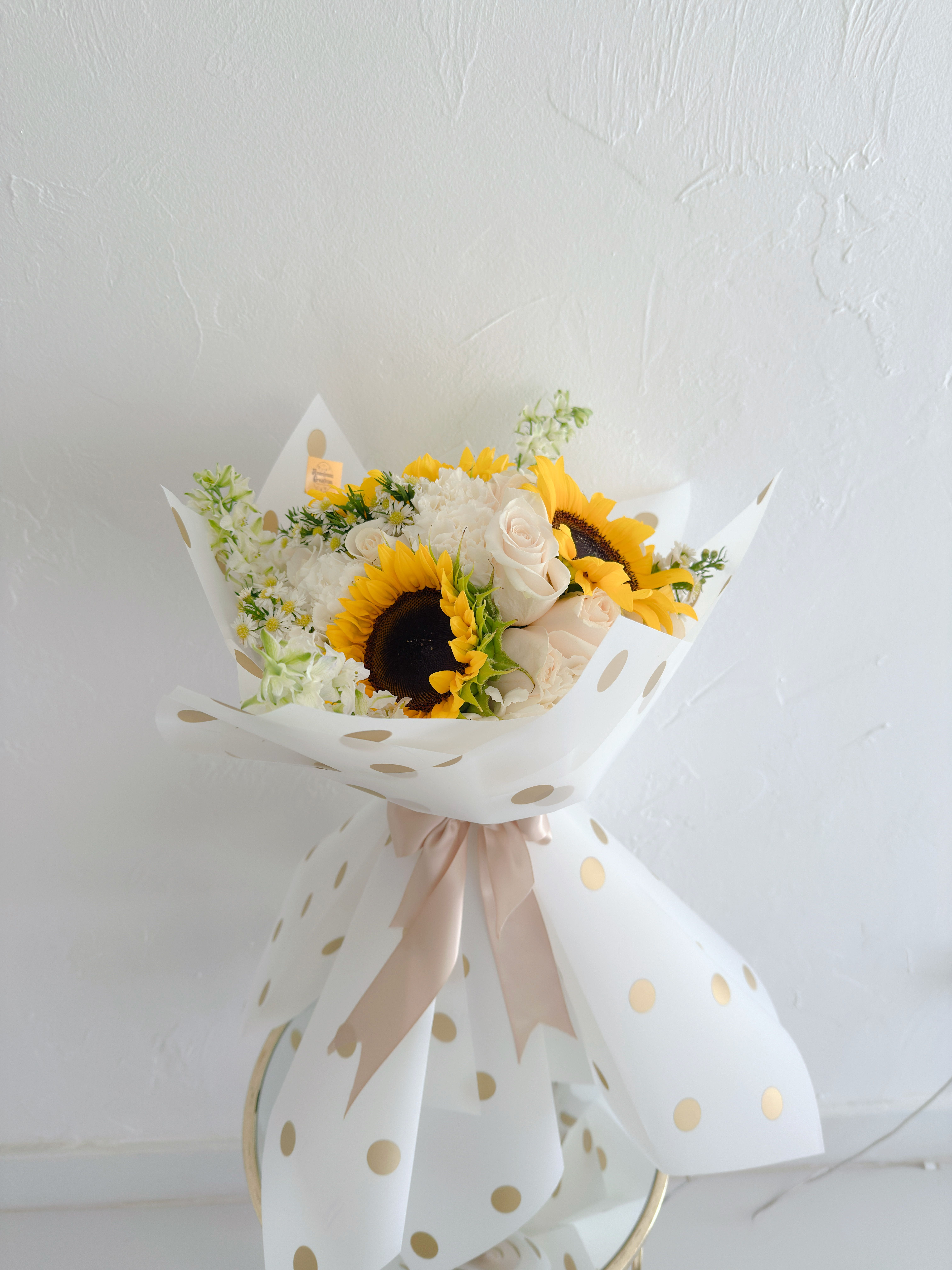 Bouquet Sun  - Bright and lively bouquet featuring vibrant sunflowers as the focal point, complemented by white roses and delicate filler flowers in soft tones. Wrapped in premium white paper with gold details and finished with a satin ribbon, this design radiates joy, warmth, and elegance.  Perfect for celebrating special moments, surprising someone with a cheerful gift, or brightening any space with its natural charm. 