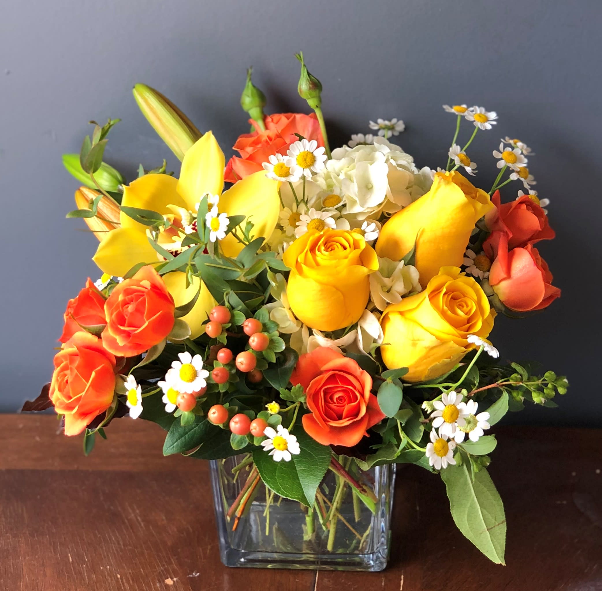 Bright arrangement of yellow roses, orange roses, lilies, and white daisies in a clear glass cube vase