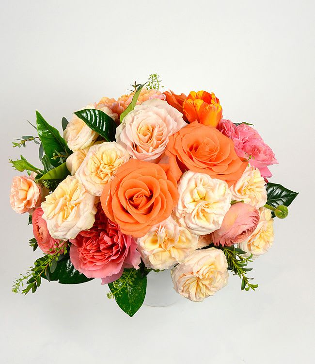 Fresh Citrus - A lush, rounded bouquet of warm pastels designed to make the person you're thinking of feel cherished - perfect as a birthday bouquet, anniversary flowers, or a Mother's Day gift. This flower arrangement brings together spray roses, garden roses and classic roses in peach, blush, pink and cream tones, with bright orange accents that keep the look fresh and cheerful for spring or any season.  The blooms are gathered tightly into a full, domed shape with ruffled carnations woven through for added texture and volume. Glossy dark green foliage peeks out around the edges, creating a striking contrast that lets the soft petals sing. Arranged in a simple white vase, the composition reads as elegant yet approachable and will fill a room with a soft, romantic presence.  Send this as a thoughtful thank-you, a just-because surprise, or a romantic gesture - she'll feel genuinely seen when presented with these abundant, garden-fresh roses and carnations. Standard vase sizing fits most tabletops; to keep blooms looking their best, refresh the water every few days and trim stems at an angle when you change it.
