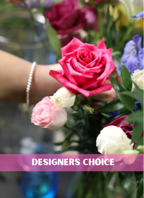 Florist hand holding a bright pink rose with small white and blush roses, labeled designers choice