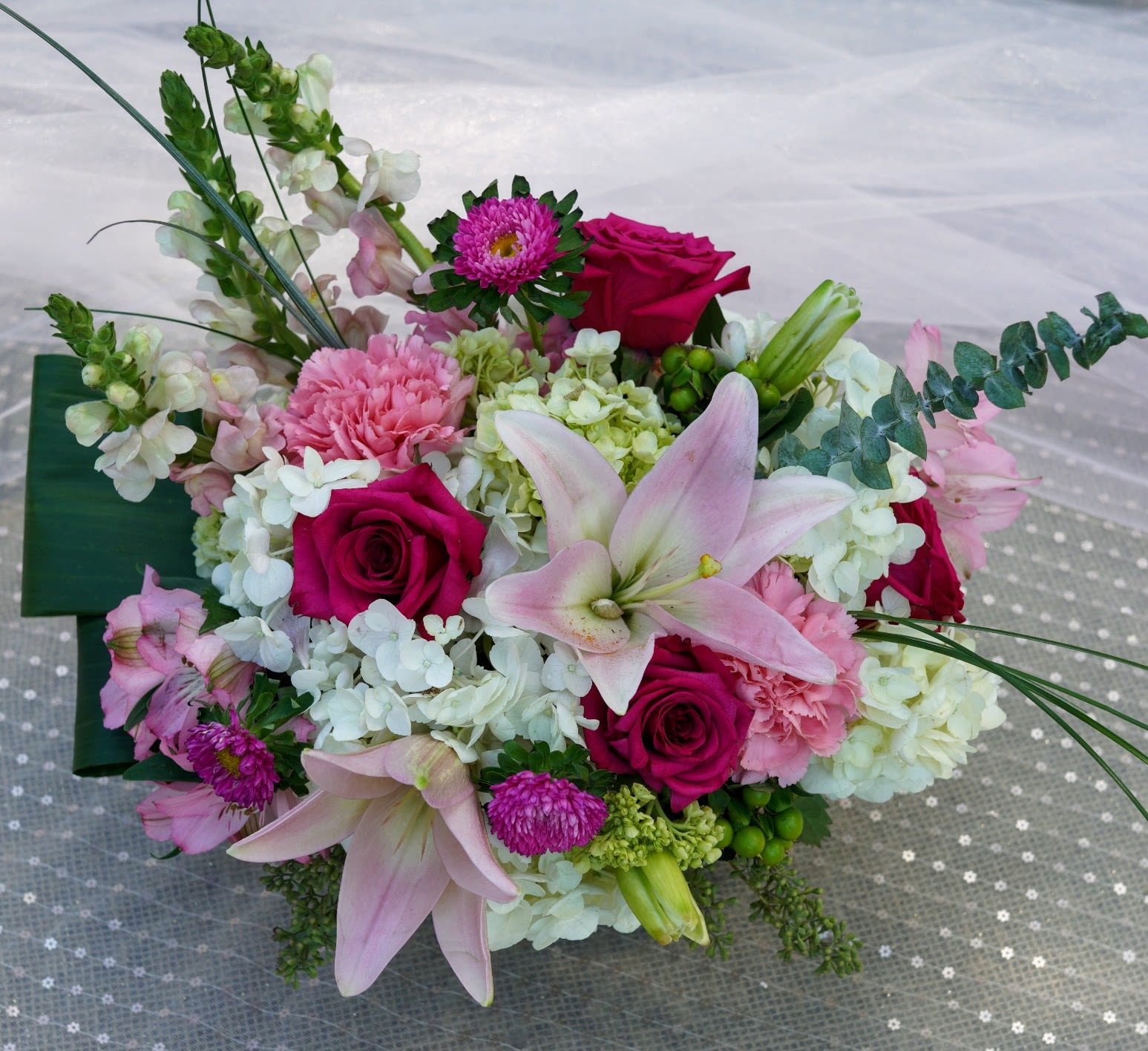 Mary Ellen's Expression - Large pale pink lilies open above hot pink roses and white hydrangea clusters to create a romantic, celebratory flower arrangement perfect for birthdays, anniversaries, or "just because" moments. The mix of lilies, roses, hydrangeas, carnations, daisies, and alstroemeria gives the person you're thinking of a lush, layered bouquet that reads both fresh and full-she'll feel genuinely celebrated the moment it arrives.  Soft pink carnations and airy white blooms balance the vivid pinks while small purple daisy-like flowers add a playful contrast against dark green foliage. Finished with a dark green decorative wrap for a clean, modern base, this design sits comfortably on a table or mantel and looks abundant from every angle. Order it to brighten someone's day or mark a special moment-arrives ready to display and enjoyed for days with regular fresh water and a cool location away from direct sun.