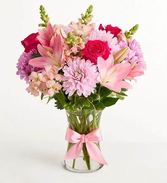 Pink Splendor - Bright pink roses and pale pink lilies mingle with lavender chrysanthemums in a soft, rounded bouquet that immediately feels like a celebration - ideal as a housewarming flower arrangement or a joyful new baby gift. Pink Splendor's pastel base is lifted by a few vivid red focal blooms and touches of purple and white, so the person you're thinking of will feel both serenely comforted and delightfully noticed the moment they see it.  This arrangement pairs roses, lilies, chrysanthemums, alstroemeria, and snapdragons in a clear glass vase finished with a pink satin ribbon for a polished, gift‑ready presentation. The full, rounded shape and mix of textures - velvety rose petals, trumpet lilies, feathery mums, and vertical snapdragons - create visual depth while remaining easygoing and fresh. Colors range from soft blush and lavender through bright pink to accents of red and purple, making it versatile for many tastes.  Perfect for saying "thank you," welcoming a newborn, or brightening Women's Day, Best Friends Day, Admin Professionals' Week, Parent's Day, a quinceañera, or Valentine's Day, this bouquet reads as thoughtful and celebratory without feeling seasonal. Arrives arranged in the vase shown and ready to display; to keep blooms looking their best, change the water every 2-3 days and trim stems slightly when you refresh the arrangement.