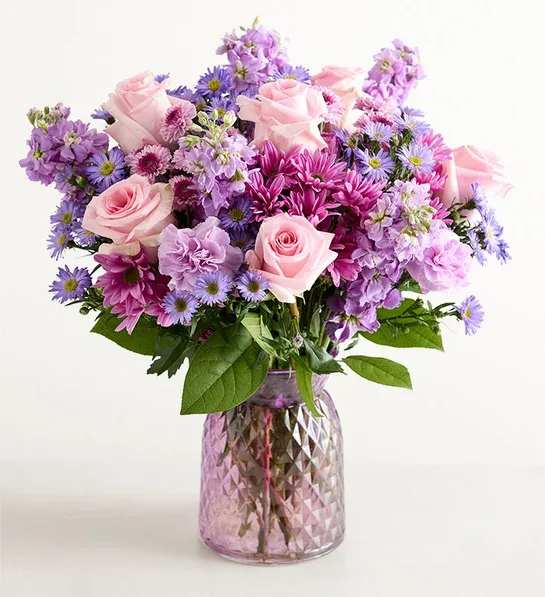 Lavender Haze - Pale pink roses mingle with lavender stock, purple asters, and white chrysanthemums to create a soft, romantic flower arrangement perfect for a birthday bouquet, anniversary surprise, or a springtime gift. The rounded, tightly layered design fills the eye with pastel color and plush texture so the person you're thinking of will feel cherished the moment it arrives.  Arranged in a pink faceted glass vase, Lavender Haze has a lush, abundant silhouette that reads fresh and elegant rather than formal. The mix of roses, stock, asters, and chrysanthemums gives a variety of shapes-velvety rose petals, feathery asters, and full chrysanthemum heads-so the bouquet feels full from every angle.  This arrangement is ideal for her milestone moments or as a thoughtful "thinking of you" surprise; it brings a gentle, feminine charm to a bedside table or dining room centerpiece. Vase included for immediate display; keep flowers in fresh water and out of direct heat to prolong their life.