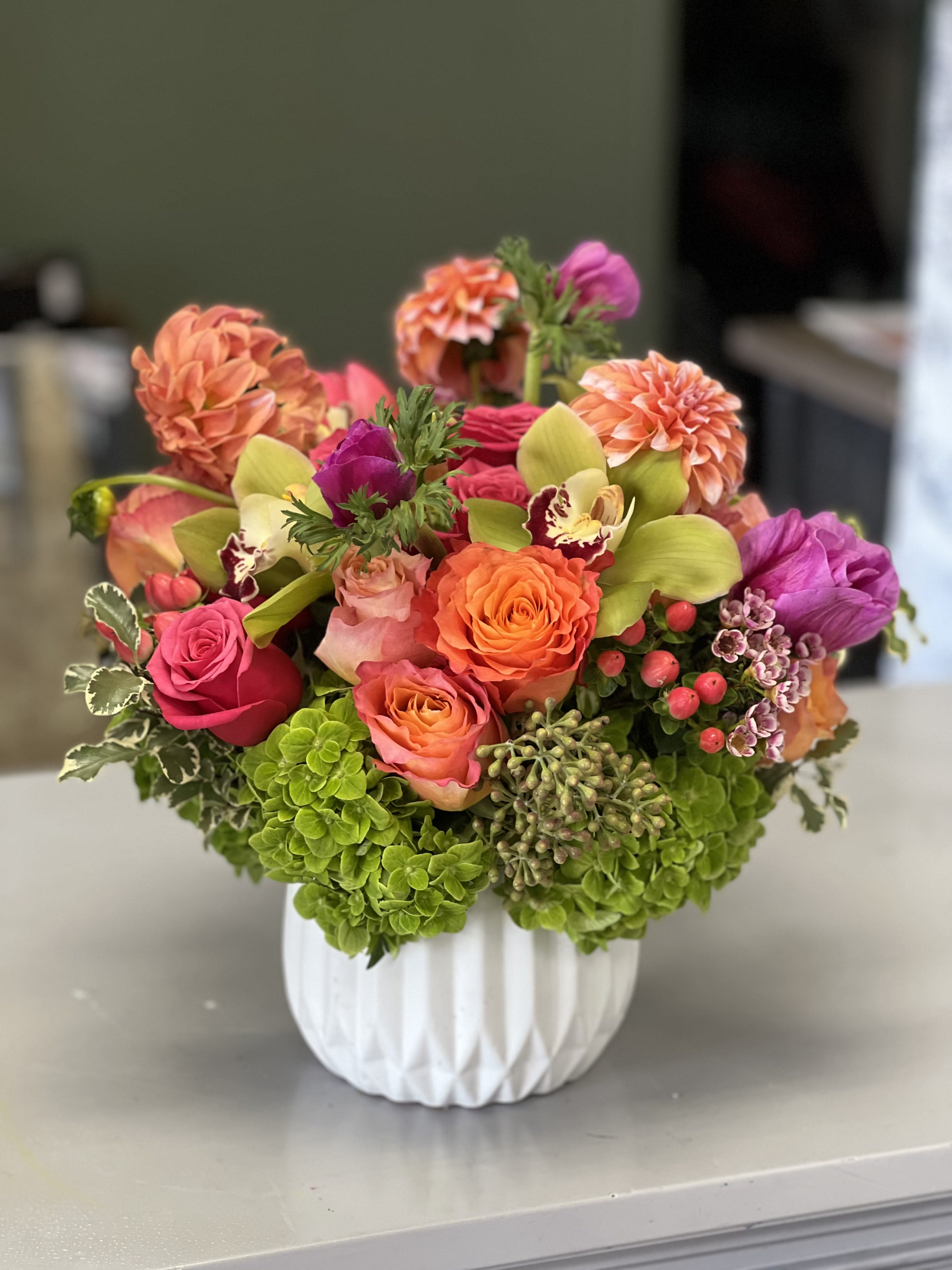 California Dreaming - This gorgeous arrangement brings all of the warmth of a California sunset! Featuring orange Dahlias and Roses, with Anemones, Hypericum, and Cymbidium Orchids, set in lush green Hydrangea in a white ceramic vase