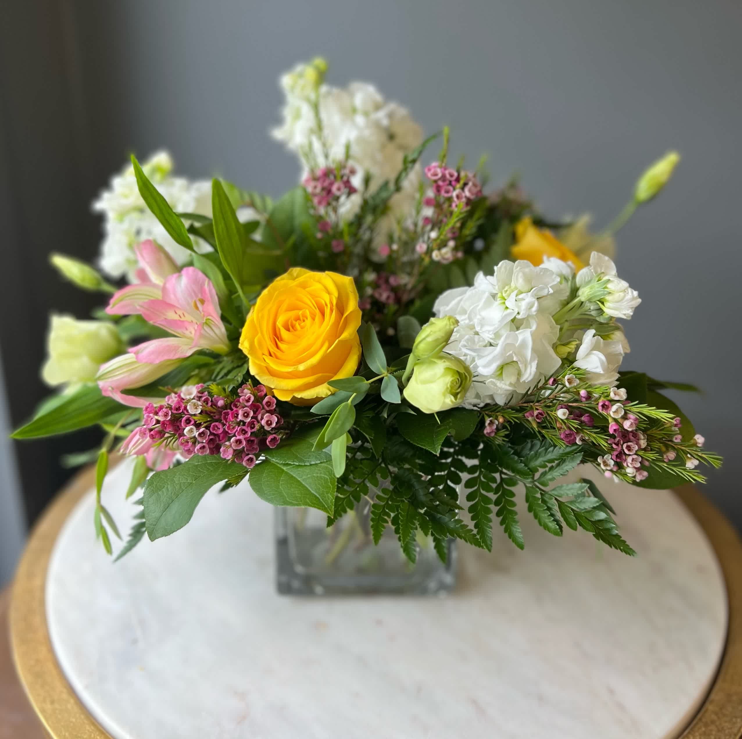 Low arrangement of yellow roses, white flowers, and pink blooms in a clear glass cube vase