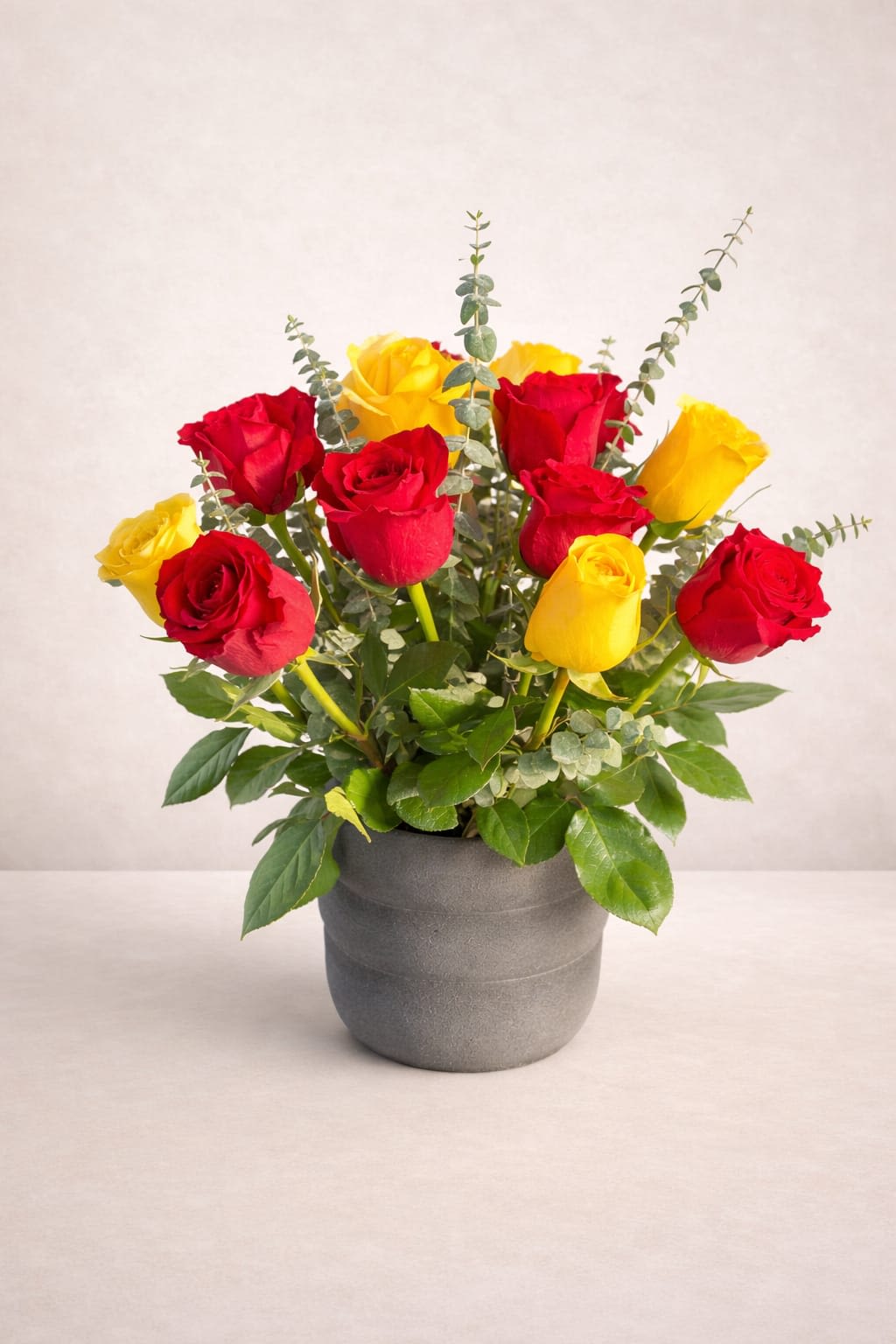 Sun &amp; Love Roses - A compact cluster of red and yellow roses arranged upright in a matte gray ceramic pot - this flower arrangement is a cheerful everyday bouquet perfect for birthdays, congratulations, or a romantic gesture. The bright red and sunny yellow roses read as both joyful and classic, making the person you're thinking of feel celebrated and seen the moment it arrives.  The arrangement pairs full rose heads with airy eucalyptus stems that add height and a soft, textured contrast against the polished gray pot. Colors: red, yellow, and green; container: gray ceramic pot; style: compact and evenly spaced for a neat, modern presentation. This is an effortless gift that looks polished on a desk, entry table, or bedside - minimal care required: fresh water in the floral foam or vase and a cool spot away from direct sun will keep the blooms looking their best.