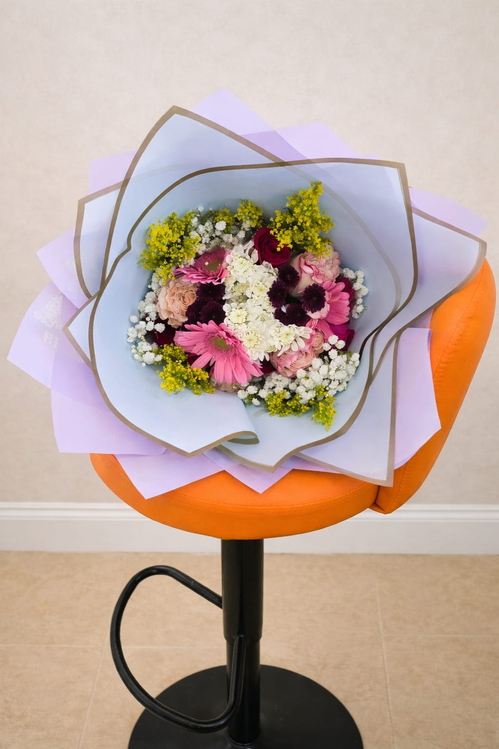 Color Burst Bouquet - A riot of pink gerbera daisies and soft pink roses makes an instant, joyful statement in this hand-tied flower arrangement - perfect as a birthday bouquet, a thank-you gift, or a just-because surprise. The rounded composition pairs bright, saturated blooms with pastel lavender and light blue wrapping paper to create a playful yet polished presentation that will make the person you're thinking of feel celebrated and seen.  This Color Burst Bouquet includes gerbera daisies, roses, carnations, chrysanthemums and other seasonal stems, accented by dark burgundy flowers and sunny yellow filler for contrast. The arrangement is finished in paper sleeves and a bouquet wrap for easy gifting; the mix of clustered white blooms and lavender-toned packaging gives the bouquet a fresh, contemporary look that suits a wide range of recipients and occasions.