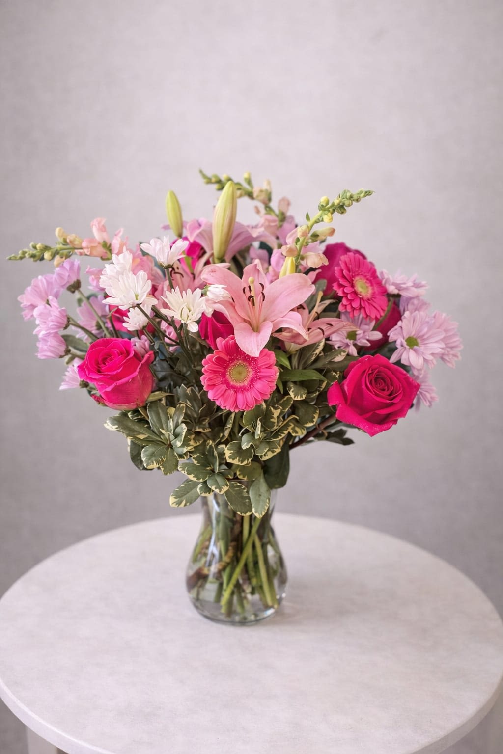 Pink Whisper Bloom - Bright pink roses and hot pink gerbera daisies take center stage in this everyday bouquet, making Pink Whisper Bloom an ideal birthday bouquet, thank-you arrangement, or "just because" surprise for someone who loves feminine, cheerful florals. The mix of pale pink lilies and lavender-and-white chrysanthemums adds soft romantic depth, while variegated greenery gives the design a fresh, garden-picked feel-she'll feel noticed and celebrated the moment she sees it.  This floral arrangement pairs roses, lilies, gerbera daisies, chrysanthemums, and snapdragons in a full, rounded shape set in a clear glass vase. The layered textures-from the silky lily petals to the bold daisy centers and delicate chrysanthemums-create lively movement and visual contrast in pink, white, purple, and green. Sized to make an uplifting impression without overwhelming a tabletop, it arrives ready to display and brighten a home or office.  Perfect for moms, friends, coworkers, or anyone who appreciates bright, feminine color, Pink Whisper Bloom sends a warm, thoughtful message without saying a word. For best enjoyment, keep the vase topped up with fresh water and trim stems every few days to extend bloom life.