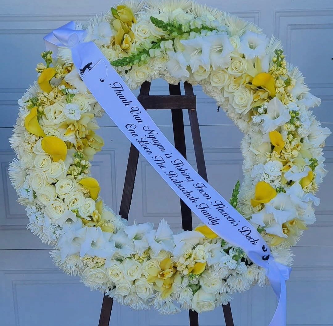 Golden Light Remembrance Round Wreath - A luminous circle of white roses and chrysanthemums forms the calm, respectful centerpiece of this sympathy wreath - a fitting remembrance tribute for a funeral or memorial service. Bright yellow calla lilies  woven through the ring to add gentle warmth and uplift the arrangement, while a lmemorial ribbon drapes diagonally across the wreath to carry a printed message of condolence.  Designed to honor and comfort the person you're thinking of, this wreath presents a dignified expression of sympathy at a service or graveside. It arrives ready to display on the easel with the memorial ribbon in place; if you'd like a specific message on the ribbon, include wording at checkout so we can personalize it for the family.