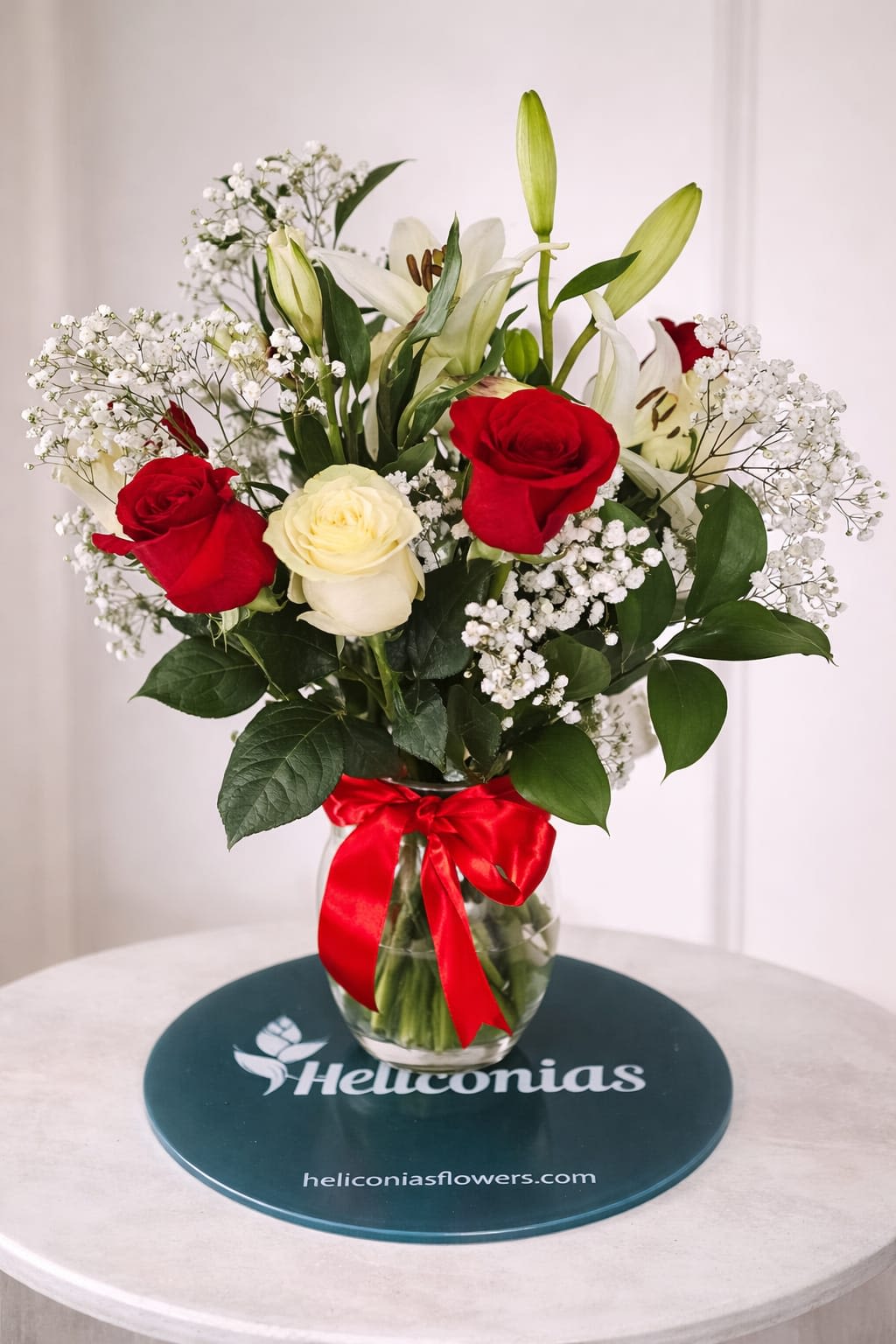 Classic Love Bloom - Red and ivory roses paired with white lilies create a classic romantic statement-perfect as a birthday bouquet, anniversary flowers, or a Valentine's Day gift to make someone feel deeply cherished. The upright composition balances tall, fragrant lilies with a dense central cluster of roses so the recipient sees both elegance and abundance at first glance.  This arrangement also includes delicate sprays of baby's breath for a soft, cloud-like texture and rich greenery that frames the blooms. Delivered in a clear glass vase and finished with a bright red satin ribbon, it arrives ready to display on a dining table or bedside stand. The polished presentation and timeless red, white, and green palette make this a memorable way to celebrate love or mark a special milestone.