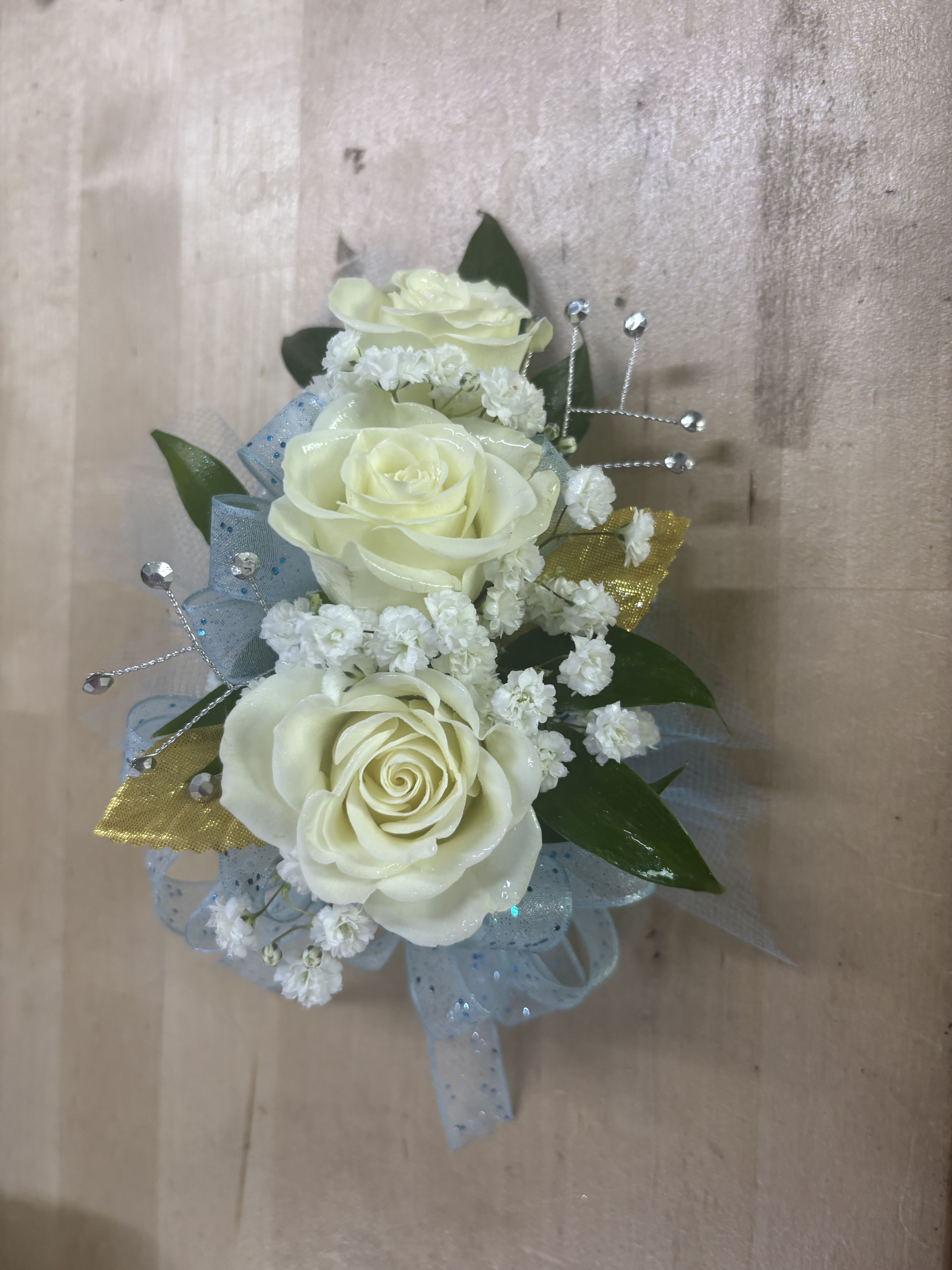 White, Light Blue, and Gold Rose corsage  - This classic wrist corsage features soft white roses, delicate baby’s breath, and fresh greenery, accented with sheer blue ribbon and subtle sparkle for a polished finish. Designed for prom, weddings, and formal events, it offers a timeless look that complements a wide range of styles and color palettes.  Each corsage is hand-crafted by a professional florist with attention to balance, proportion, and secure construction, ensuring it stays in place comfortably throughout the event. The combination of premium blooms and lightweight design creates a piece that is both elegant and easy to wear.  Perfect for prom, homecoming, or special occasions, this wrist corsage delivers a refined floral accent with dependable quality and a clean, finished look.