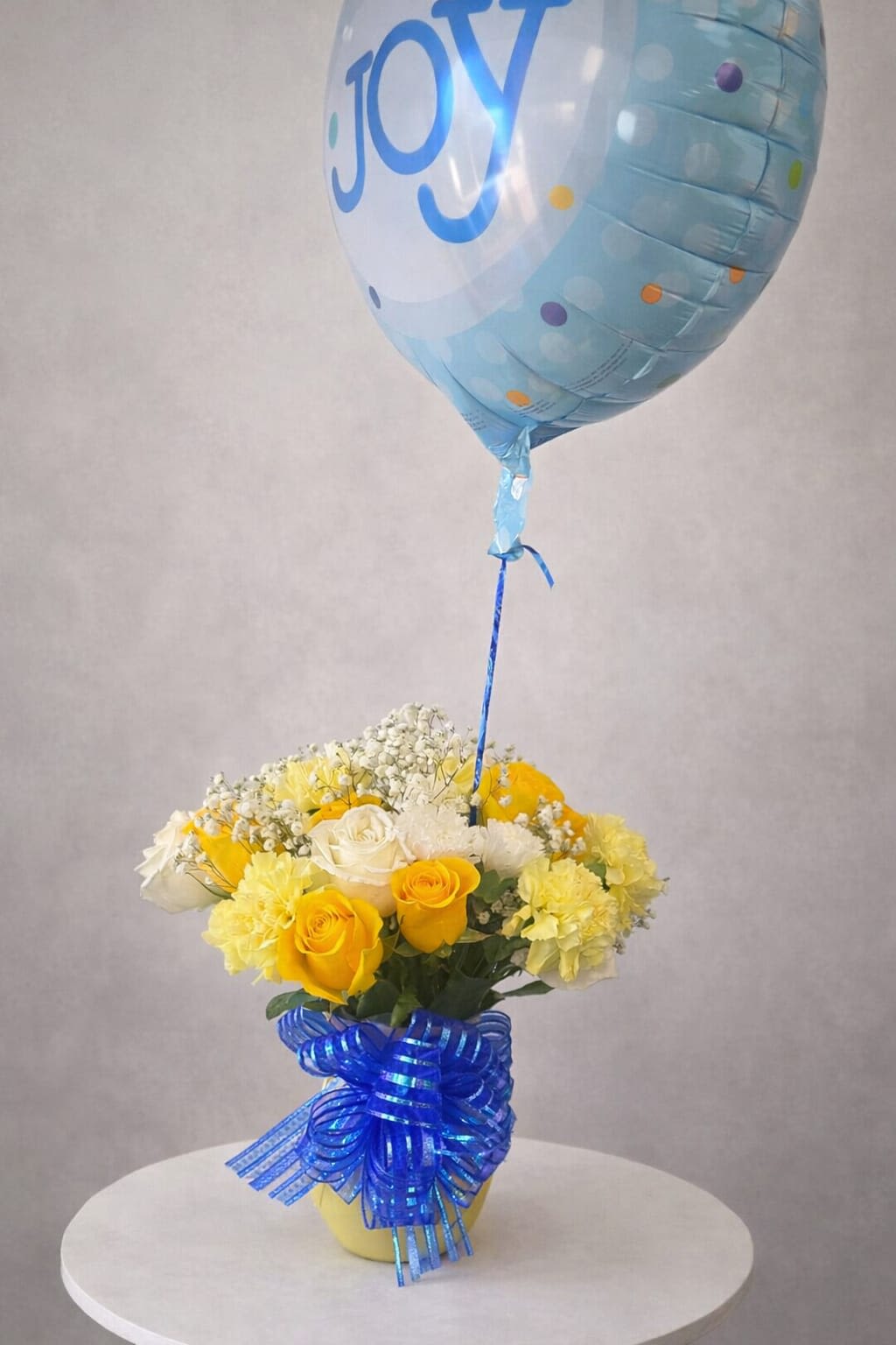 Baby Sunshine &amp; Blue - Sunlight yellow roses and pale yellow carnations arranged with crisp white filler flowers create a cheerful, compact flower arrangement perfect for a birthday bouquet, congratulations gift, or a just-because surprise. A large light-blue foil balloon printed with "Joy" rises above the flowers, tied with a blue ribbon - a playful topper that makes the recipient feel celebrated from the moment they see it.  Set in a bright yellow container, the mix of yellow, white, and blue reads joyful and uplifting without being over the top. The arrangement is intentionally compact for easy display on a table or desk; the combination of soft rose petals, ruffled carnation texture, and delicate white accents offers both visual contrast and a gentle, happy fragrance. Ideal for welcoming a new baby, brightening someone's day, or marking a small victory - it's the kind of gift that says you're thinking of them with warmth and delight.