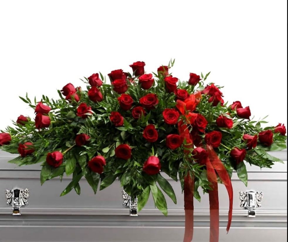 Red Roses Casket Spray  - A classic cascade of deep red roses arranged in a large, low casket spray creates a solemn, romantic tribute for a funeral or memorial service. This sympathy arrangement rests elegantly atop a silver casket, the dense clusters of velvety red roses spilling outward into abundant mixed greenery for a formal, traditional silhouette. Long red ribbon tails drape from the front, adding a restrained, honoring flourish that reads both respectful and heartfelt.  Made to cover the length of a casket, the spray's broad, symmetrical shape and generous greenery provide a dignified presence that will make the person you're honoring feel truly remembered. The red-and-green palette reads timeless and comforting; suitable year-round for a funeral or memorial. Care: designed for placement on a casket and delivered ready to display; order soon to ensure timely delivery for services.