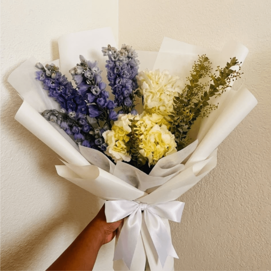 Serenity Garden - Gentle, reflective, and calm—Serenity Garden feels like a quiet walk through a blooming meadow. Soft blue delphinium and pristine white hydrangeas offer a moment of beauty and quiet support.  This seasonal bouquet features:  White Hydrangea  Light Blue Belladonna Delphinium  Fresh Boxwood  Garden Greens  Presentation: Expertly wrapped in our signature textured paper and tied with satin ribbon. Prefer a vase? Add one to your cart to complete the look.  Personal Touch: We’ll include a custom card message with every order. You’ll be able to write your message at checkout.  Please Note: All flowers are subject to seasonal availability. If a specific bloom isn’t available, our designers will thoughtfully substitute with flowers in a similar shape, style, and color.  Standard size shown. Vase upgrade is optional.   Floral Care Guide  Your arrangement arrives in our signature "double wrap" and is nestled in a protective box. While the stems are secured with a wet wrap for transit they are ready for a full drink!   Unpack: Gently lift the bouquet from the box. Remove the plastic bag and wet towel from the stems. Trim &amp; Hydrate: Trim 1/2" off stems at an angle and place in a clean vase with cool water. Refreshed: Change water daily for a long-lasting bloom. 