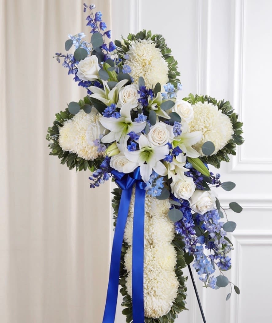 floral cross blue - A delicate standing spray  featuring crisp white roses paired with graceful blue delphinium, accented by cheerful white daisies. The soft, velvety petals of the roses create a timeless elegance, while the tall, airy delphinium adds height and a gentle pop of cool blue. Nestled throughout, the daisies bring a fresh, natural touch with their bright centers and simple beauty. Together, these blooms create a serene and uplifting display—perfect for conveying peace, admiration, and a sense of calm sophistication.