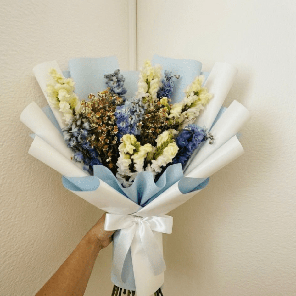 Baby Blue - Whimsical, soft, and lighthearted—Baby Blue is like a fluffy summer cloud. Fluffy white snapdragons and sapphire delphinium are paired with feverfew for a modern, sweet welcome for a new little one.  This seasonal bouquet features:  Light Blue Belladonna Delphinium  White Snapdragons  Petite White Feverfew  Garden Greens  Presentation: Expertly wrapped in our signature textured paper and tied with satin ribbon. Prefer a vase? Add one to your cart to complete the look.  Personal Touch: We’ll include a custom card message with every order. You’ll be able to write your message at checkout.  Please Note: All flowers are subject to seasonal availability. If a specific bloom isn’t available, our designers will thoughtfully substitute with flowers in a similar shape, style, and color.  Standard size shown. Vase upgrade is optional.   Floral Care Guide  Your arrangement arrives in our signature "double wrap" and is nestled in a protective box. While the stems are secured with a wet wrap for transit they are ready for a full drink!   Unpack: Gently lift the bouquet from the box. Remove the plastic bag and wet towel from the stems. Trim &amp; Hydrate: Trim 1/2" off stems at an angle and place in a clean vase with cool water. Refreshed: Change water daily for a long-lasting bloom. 