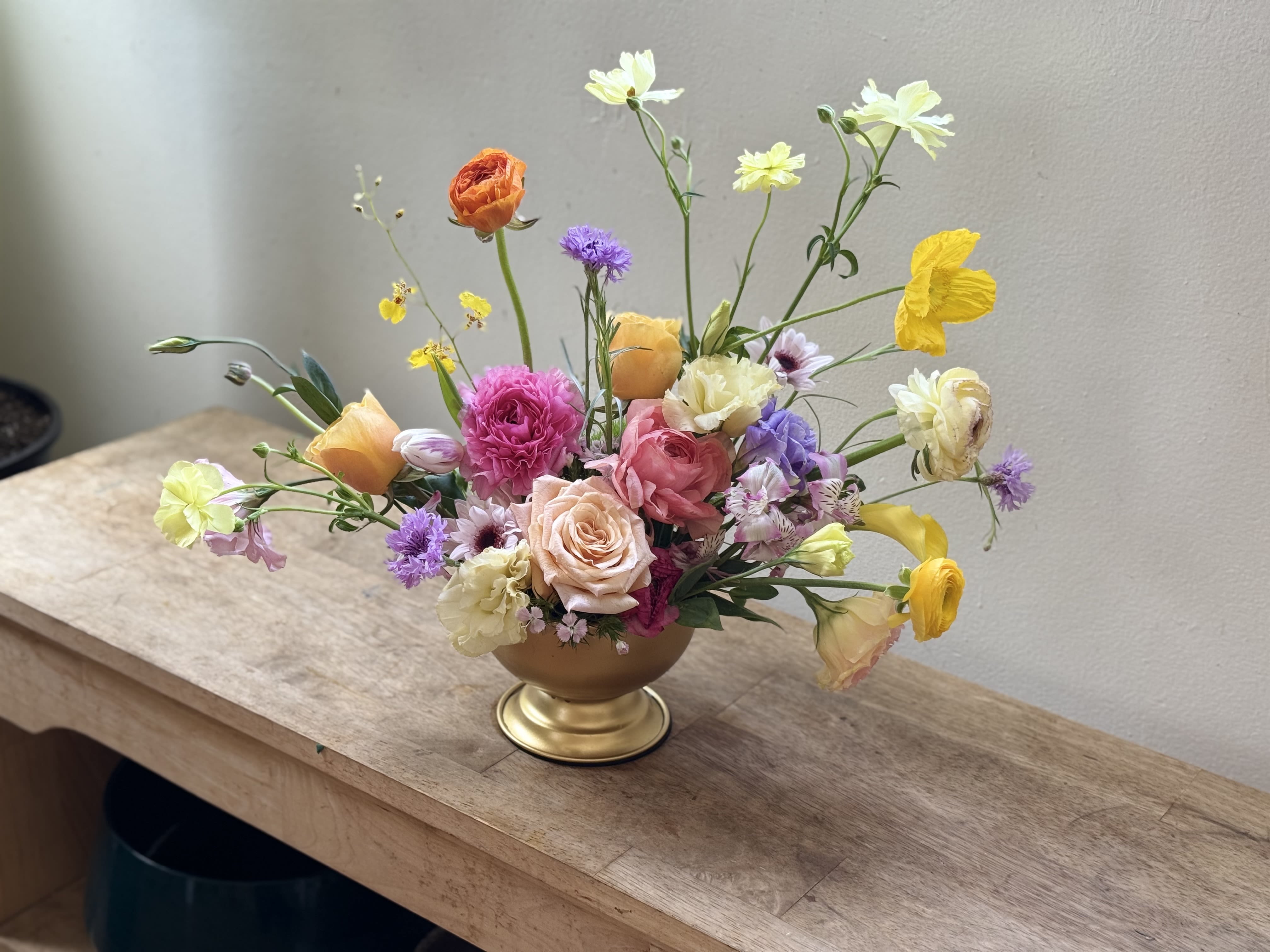 Flor - A loose, airy centerpiece of in-season blooms that greets the recipient with a bright, garden-fresh smile-perfect as a birthday bouquet, anniversary surprise, or a cheerful thank-you arrangement. Flor pairs pink and peach roses and ranunculus with ruffled carnations, lisianthus, and daisy-like blooms in a lively mix of pink, yellow, purple, white, and touches of orange to create a whimsical, celebratory feel that reads as both romantic and playful.