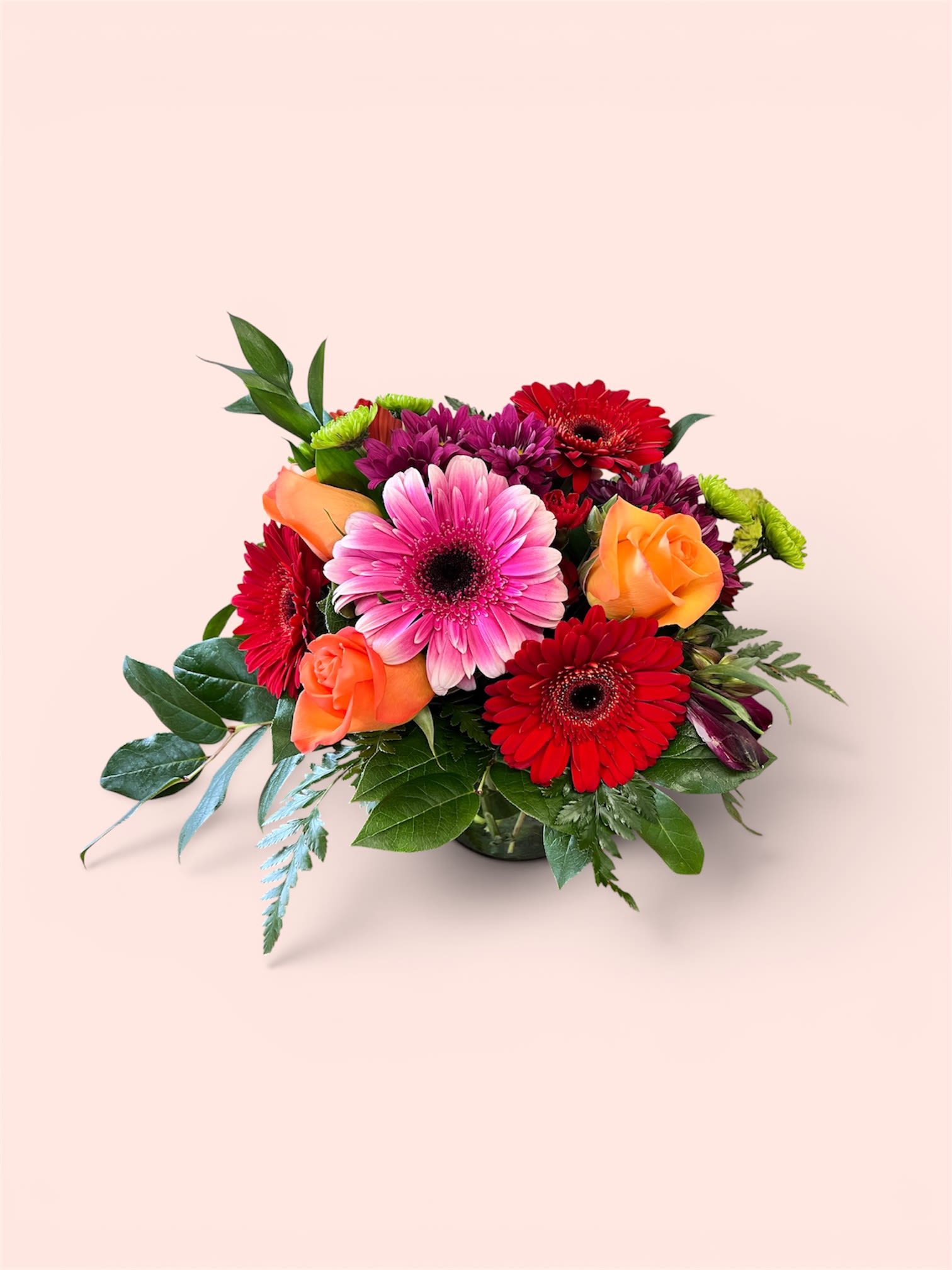 Mother’s Day Special #2 - A burst of vivid color-hot pink gerbera daisies at the center surrounded by red daisies, warm orange roses, and purple chrysanthemums-makes this Mother's Day flower arrangement an unmistakably joyful gift for mom on Mother's Day, birthdays, or to say thank you. The lively mix of gerbera daisies, roses, chrysanthemums and alstroemeria reads bright and celebratory, the kind of bouquet that will make the person you're thinking of feel seen and delighted.  Arranged tightly in a low glass vase, the design has a full, rounded silhouette with broad glossy foliage framing the blooms for extra depth and shine. Expect a cheerful palette of pink, red, orange and purple with small accent blooms woven through for texture; the compact vase makes it ideal for tabletops or a kitchen counter where she can enjoy the color up close.  Perfect for moms who love bold color and joyful arrangements, this piece suits everyday gifting, birthday celebrations, or a vibrant thank-you. Place in fresh water on arrival and trim stems if needed to refresh; available in the pictured low glass vase for immediate display.